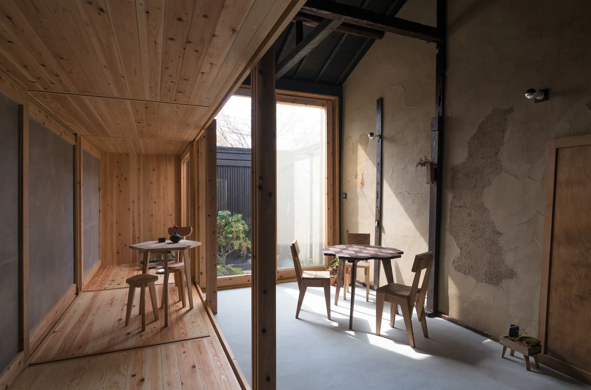 A rustic dining area features two wooden tables and chairs on a concrete floor next to a raised wood-plank platform and a raw plaster wall.