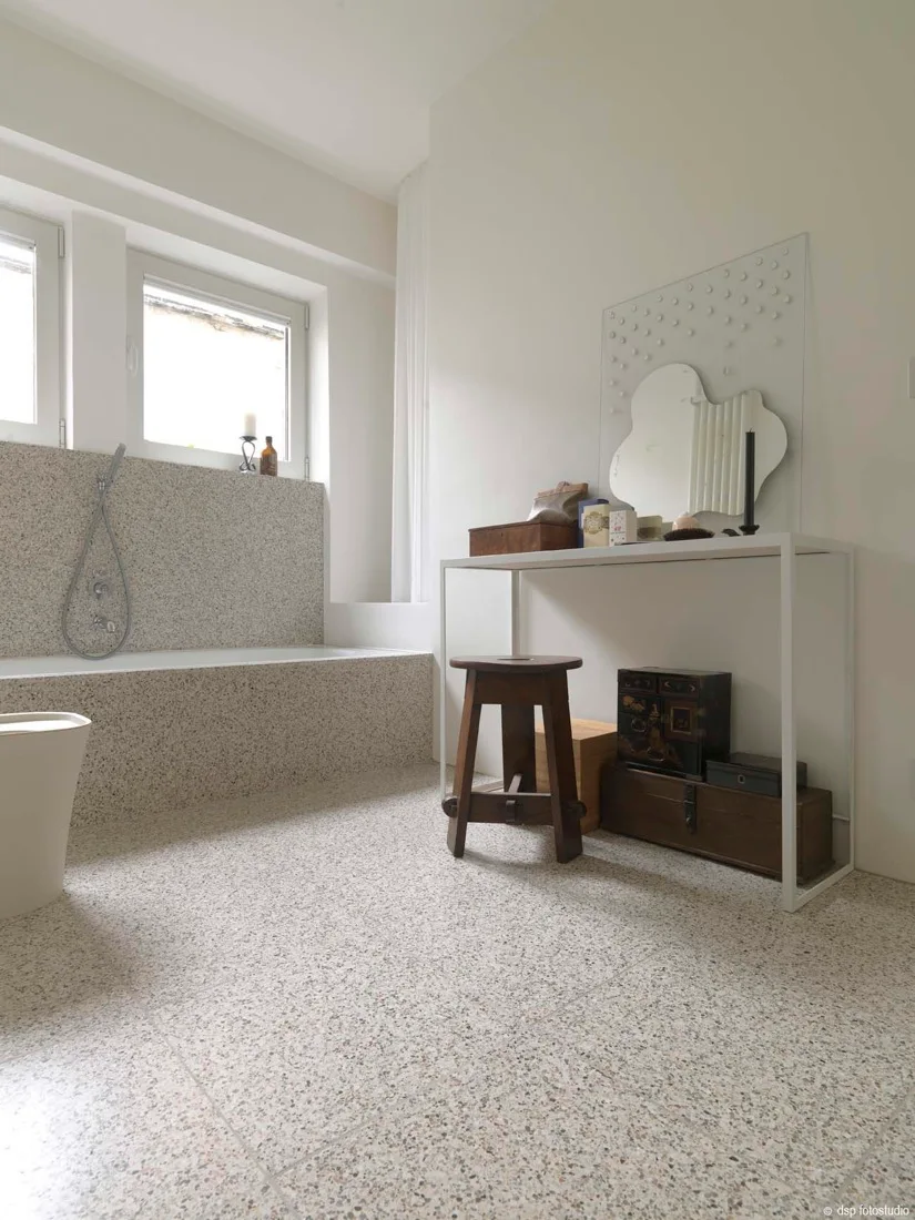 A minimalist white bathroom featuring speckled terrazzo flooring, a matching bathtub surround, a slim white console table, and a dark wooden stool.