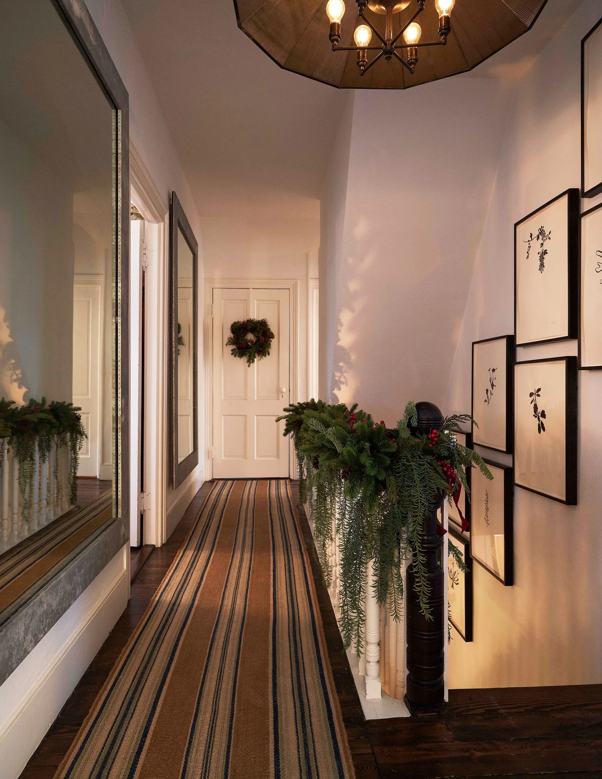 A narrow hallway features a striped runner rug, a large wall mirror, and a staircase railing decorated with festive evergreen garland.