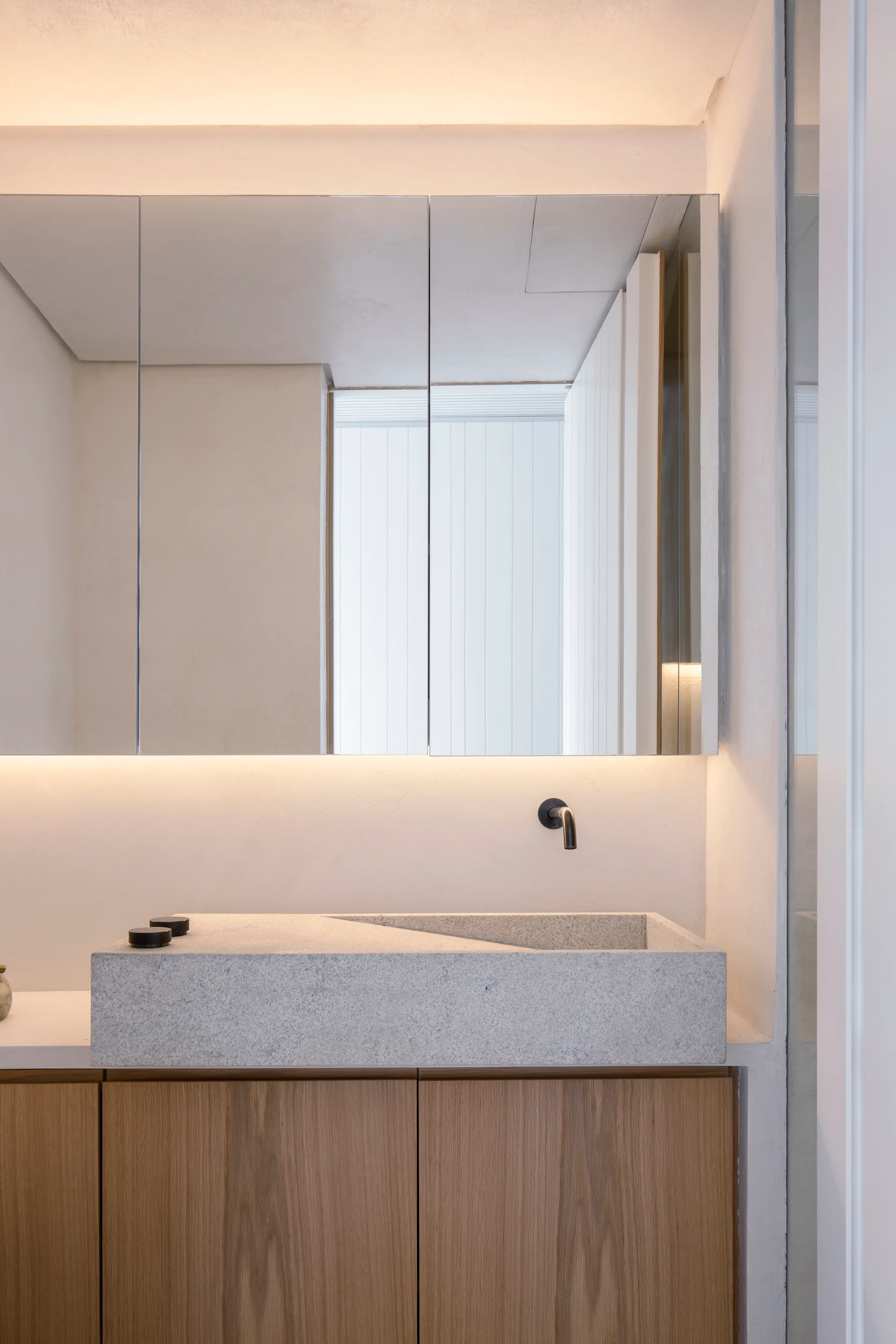 A minimalist bathroom vanity featuring a grey stone sink basin with a sloped interior, a wood cabinet, a wall-mounted faucet, and a lighted mirror.