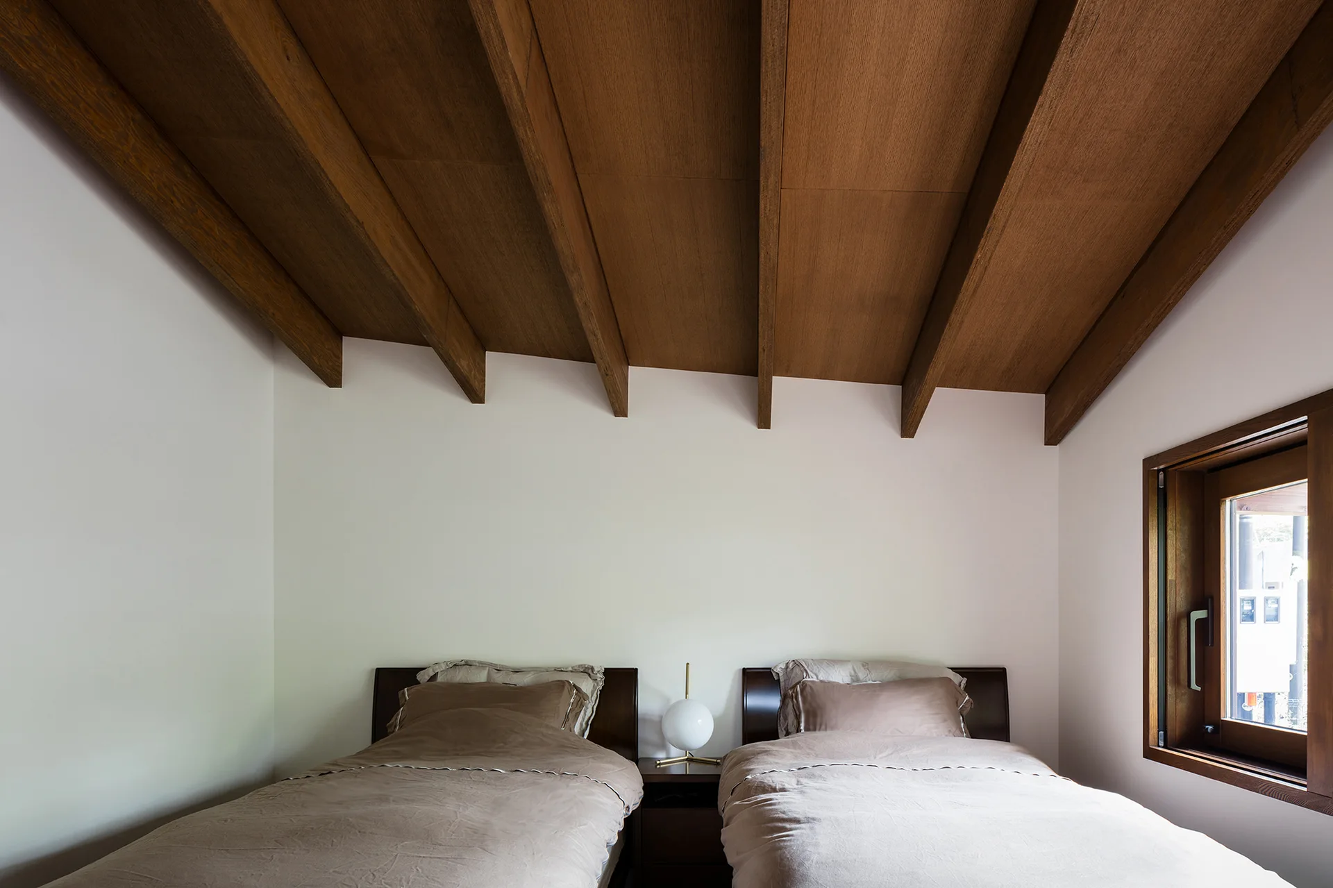 Two twin beds with brown linens sit side-by-side beneath a sloped wooden ceiling with prominent exposed beams and a central globe table lamp.