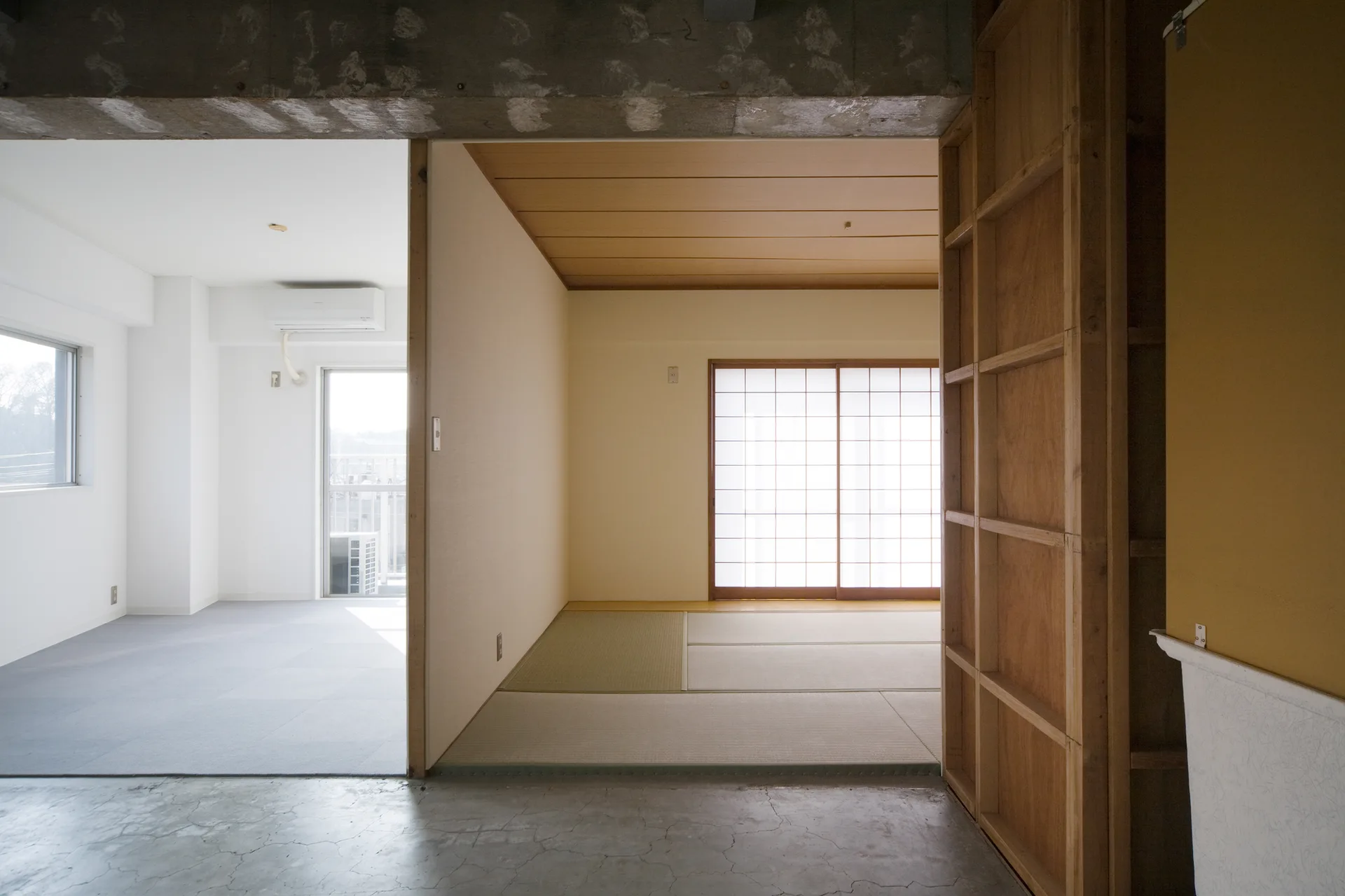 A minimalist apartment interior with a traditional tatami mat room, shoji screen doors, raw concrete beams, and exposed wood framing.