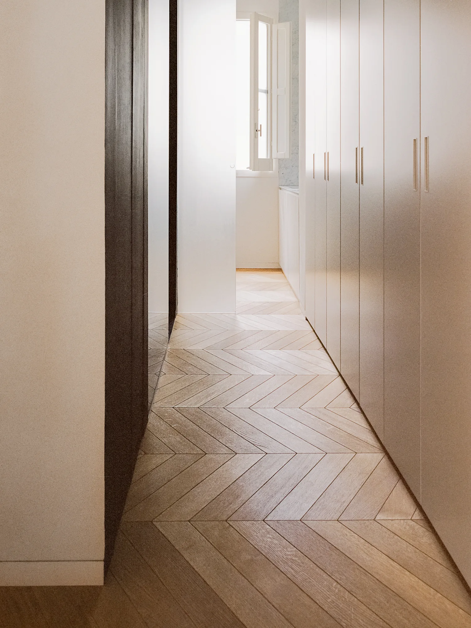 A bright hallway featuring light wood herringbone flooring, integrated floor-to-ceiling closets, and a white window at the end of the corridor.