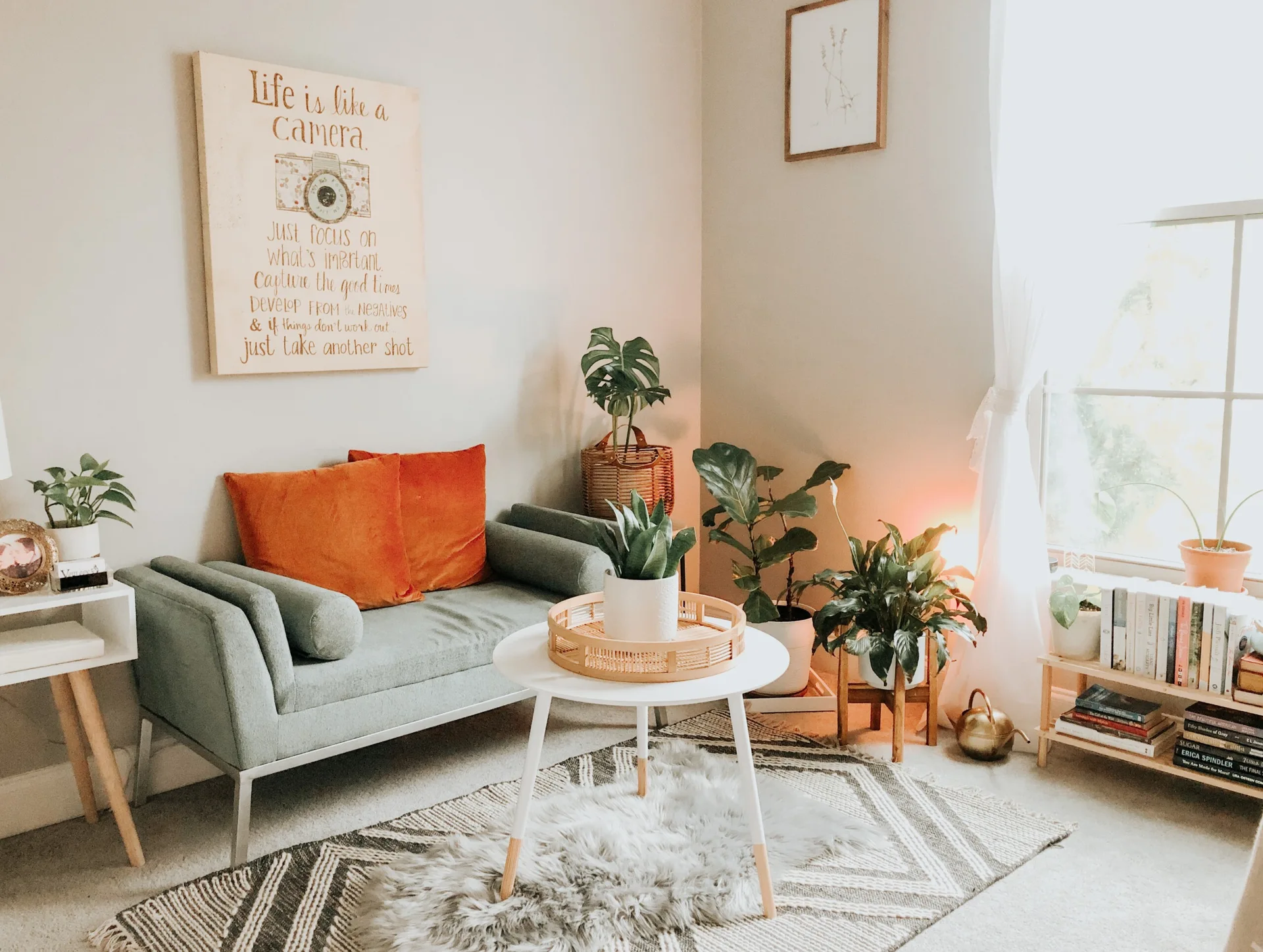 A small gray sofa decorated with bright orange velvet pillows sits in a cozy corner filled with potted green plants, layered rugs, and a coffee table.