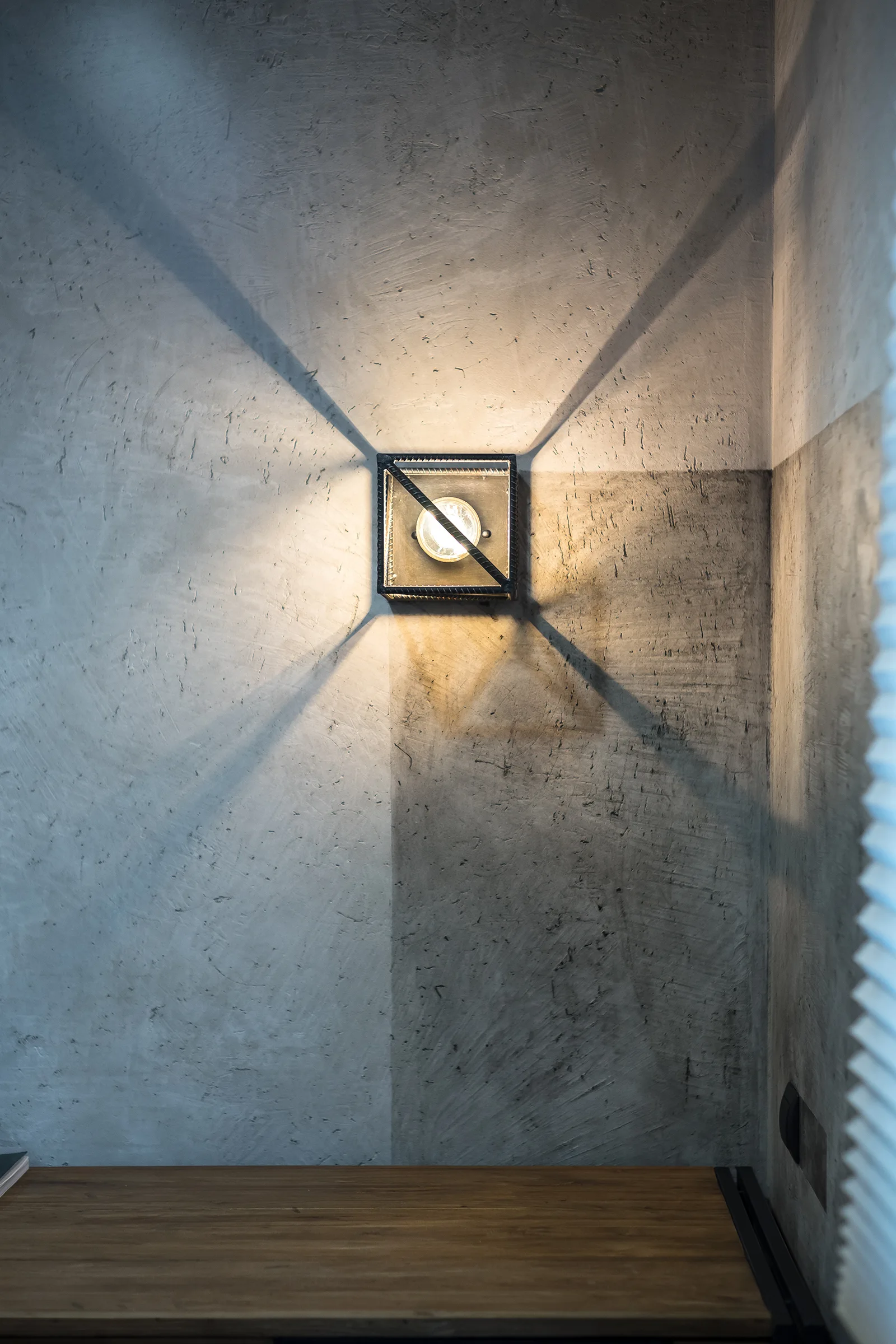A square metal wall sconce casts warm, X-shaped light beams onto a textured grey plaster wall above a wooden surface.