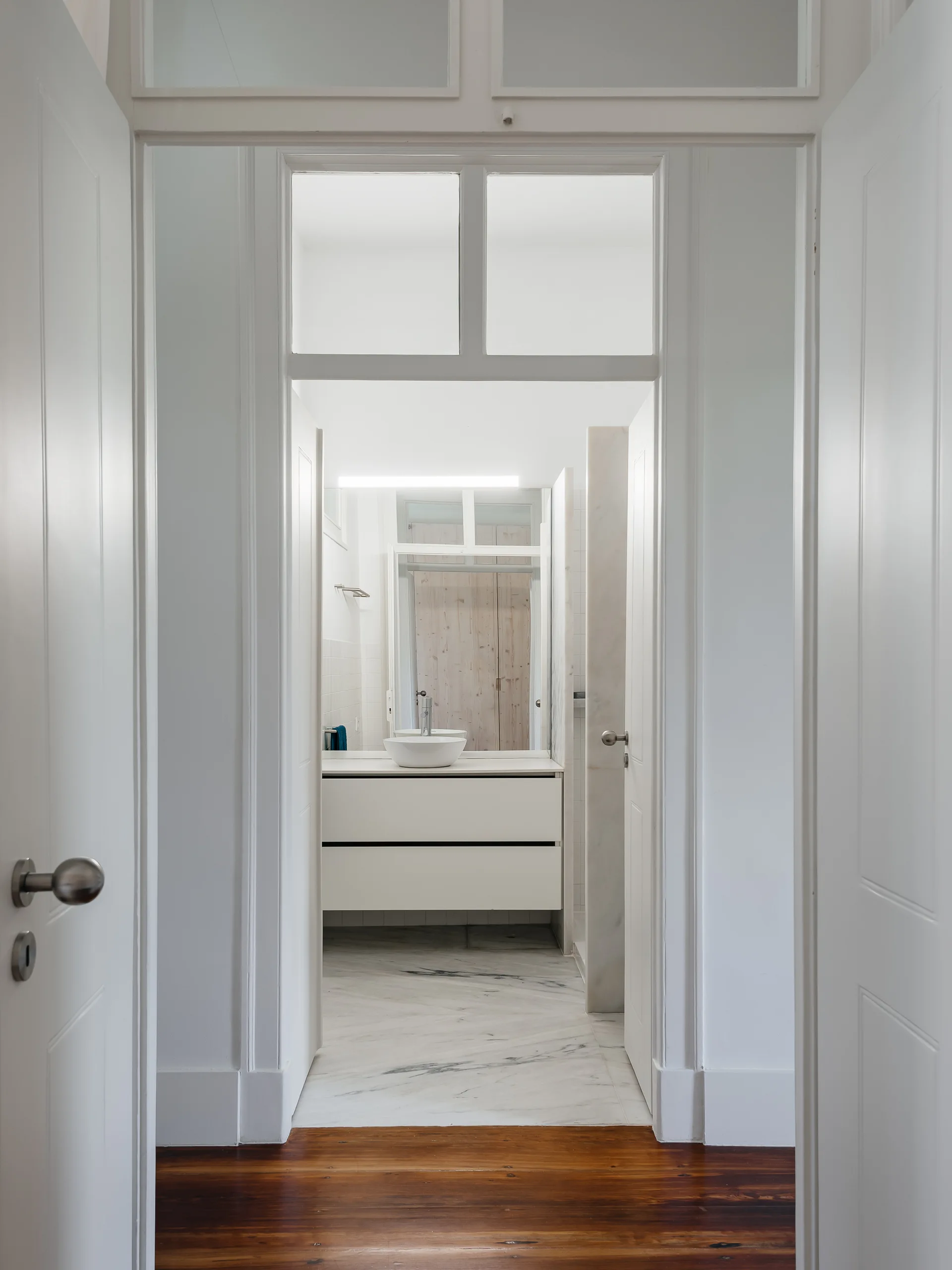 A view through a white doorway into a minimalist bathroom featuring a floating vanity, a vessel sink, and marble tile flooring.