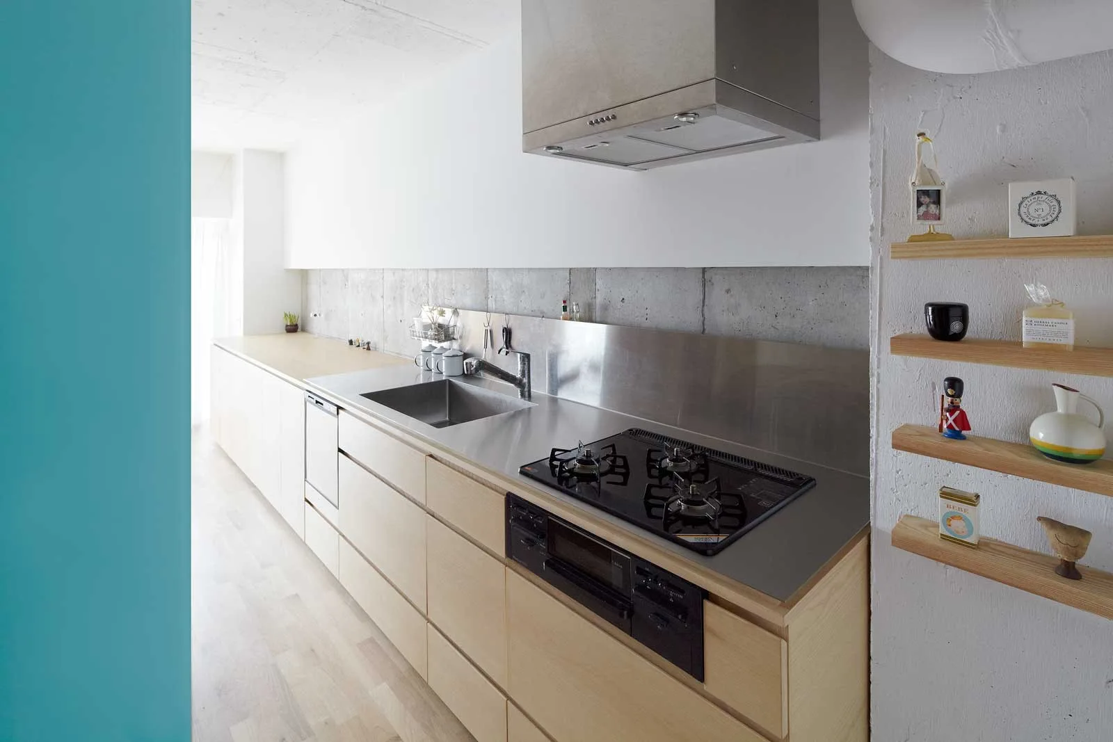 A minimalist kitchen featuring light wood flat-front cabinets, a stainless steel countertop with sink and stovetop, and an exposed concrete wall.