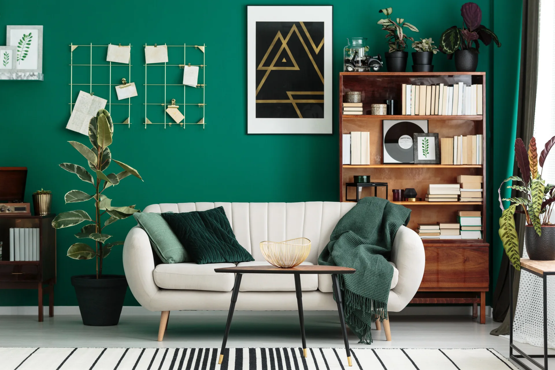 A cream-colored sofa sits against a dark green wall in a living room next to a wooden bookshelf, with a black and white striped rug on the floor.
