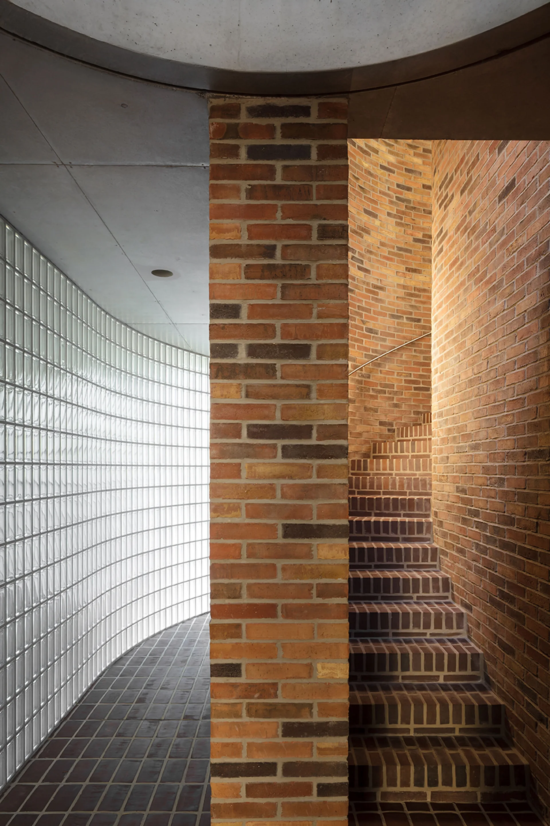A winding red brick staircase ascends next to a central brick pillar and a curved glass block wall under a concrete ceiling.