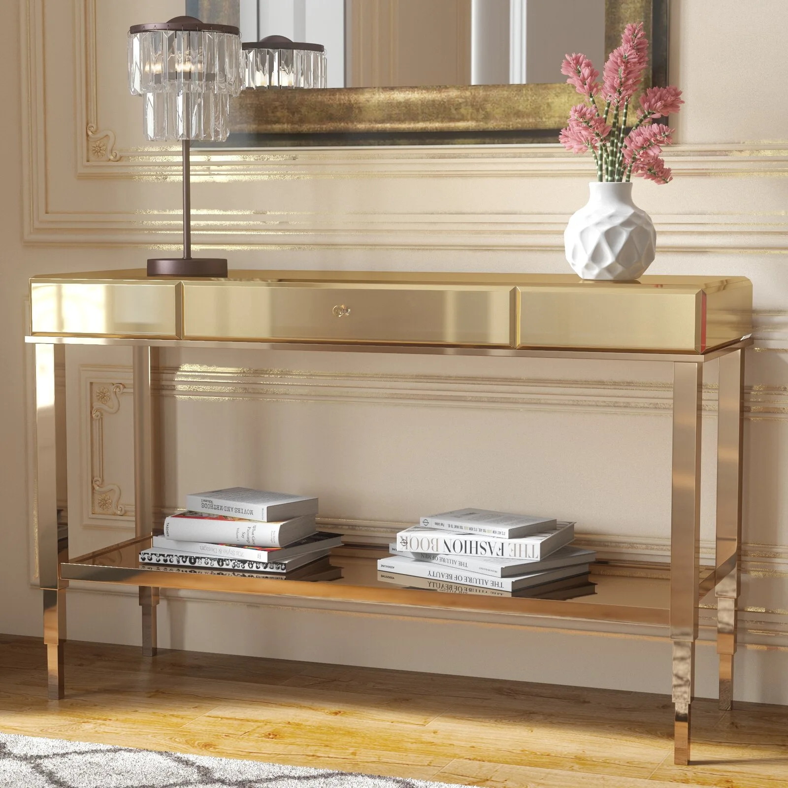 A shiny gold console table features a lower shelf with stacked books, a white vase with pink flowers, and a crystal table lamp against a paneled wall.