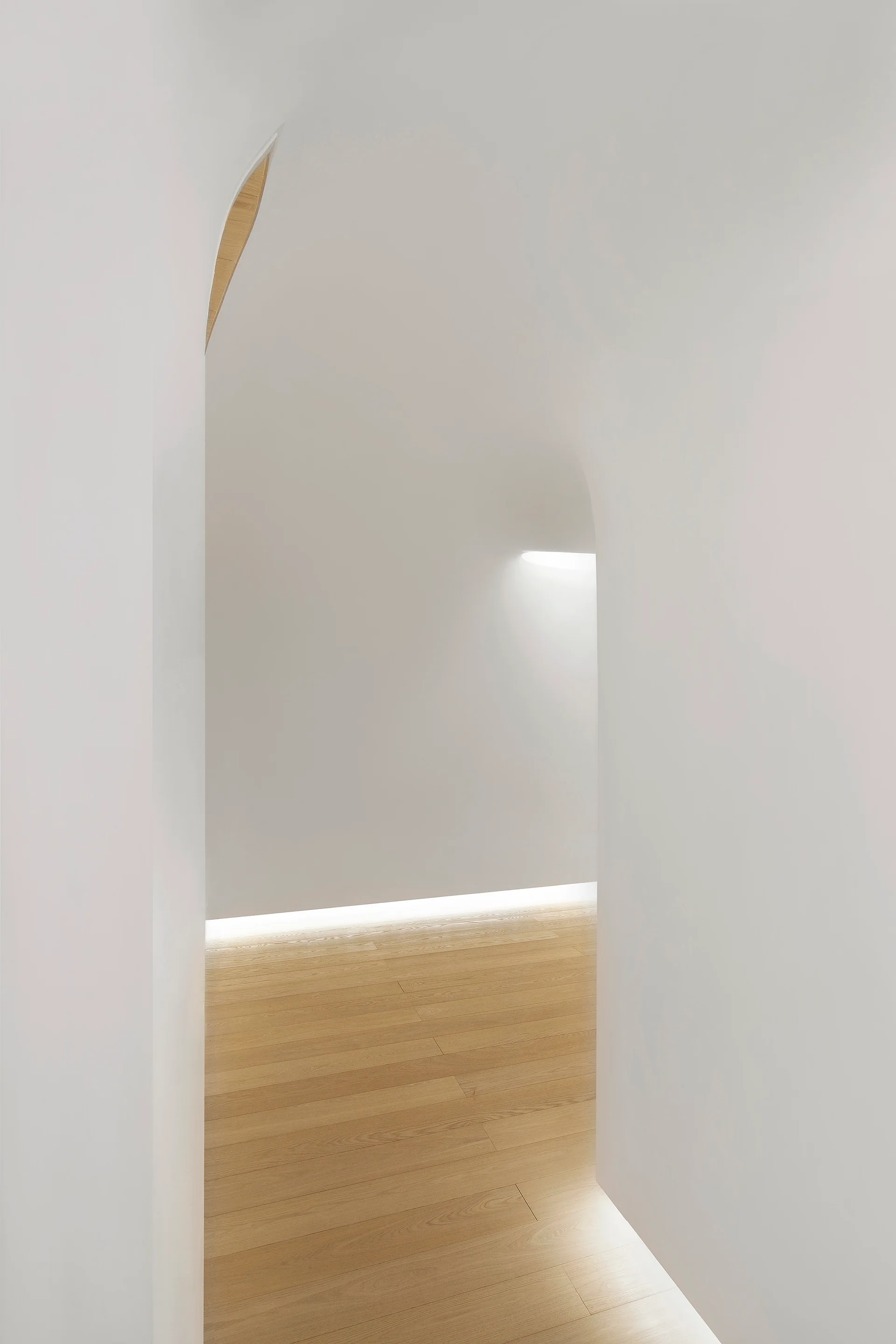 A minimalist white hallway featuring a sculptured archway, light wood plank floors, and hidden cove lighting along the floor.