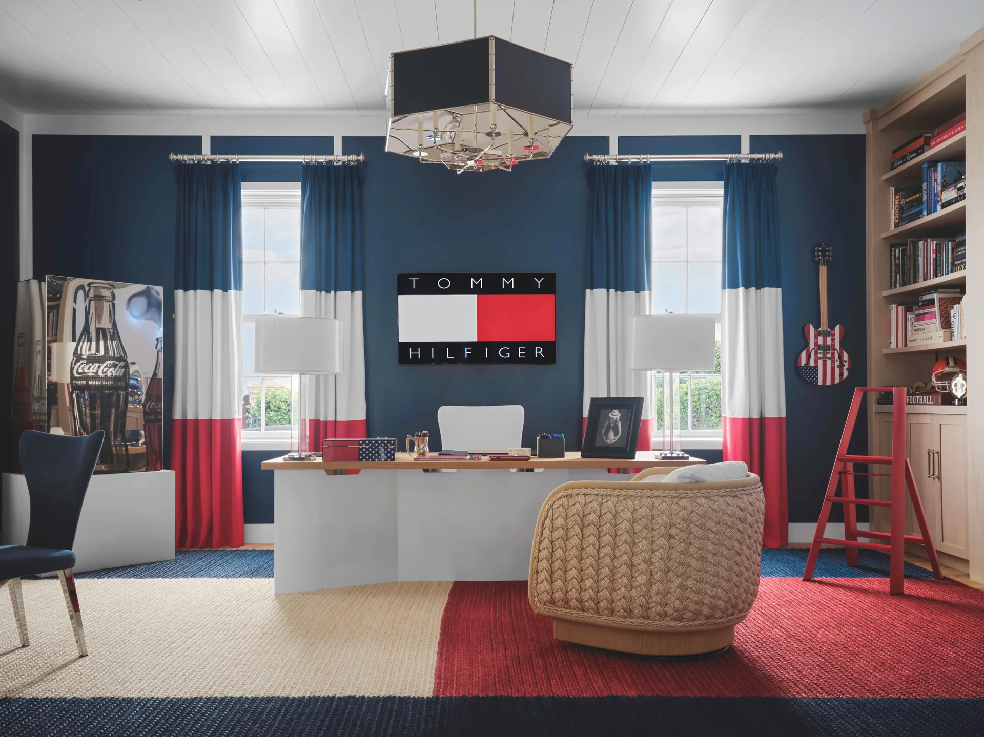 A home office featuring navy blue walls, a white desk, a textured tan woven swivel chair, and red, white, and blue color-blocked curtains.