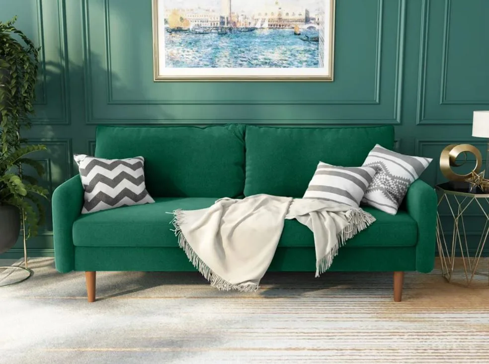 An emerald green mid-century sofa sits against a matching paneled green wall beneath a gold-framed ocean landscape painting.