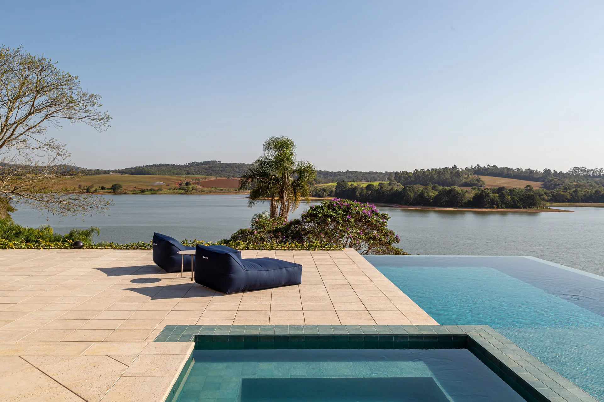 Two navy blue loungers sit on a beige stone tiled patio beside an infinity pool overlooking a scenic lake under a clear blue sky.