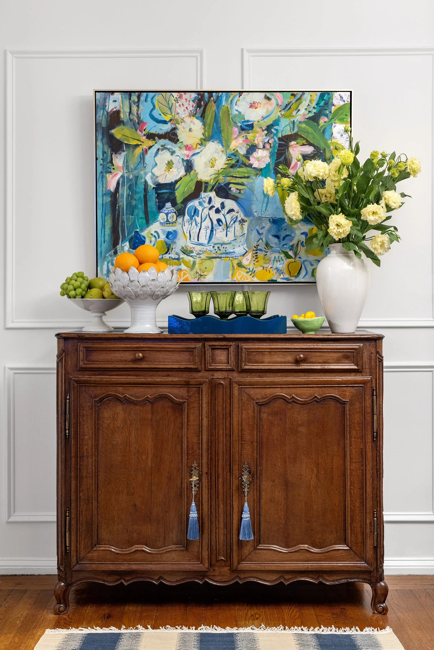 A traditional wooden credenza with tassel-keyed doors stands against white paneled walls, topped by a colorful floral painting and decorative fruit bowls.