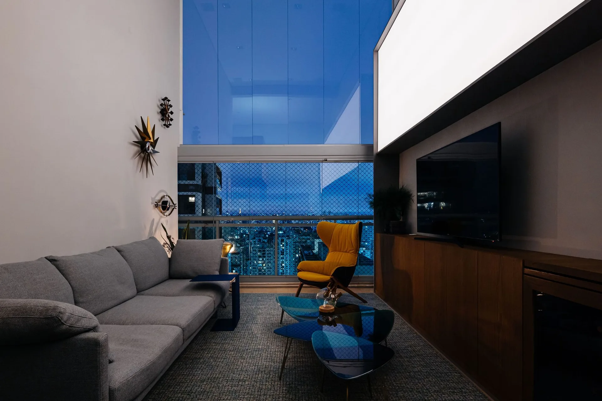 A modern living room with a gray sofa, mustard wingback chair, blue glass nesting coffee tables, and a large window showing a city view at night.