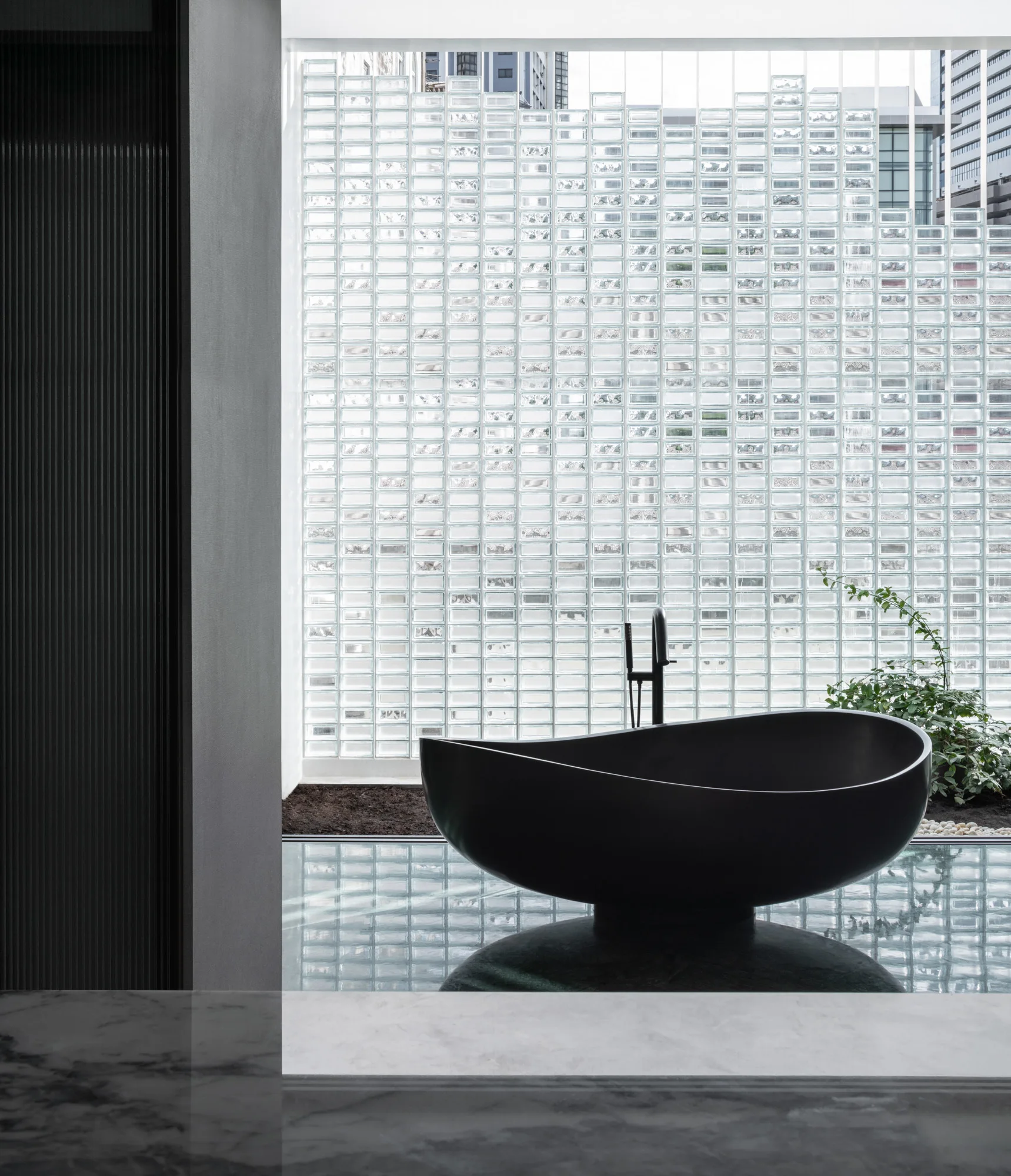 A minimalist bathroom features a black freestanding bathtub positioned in front of a translucent glass block wall and a reflective floor.