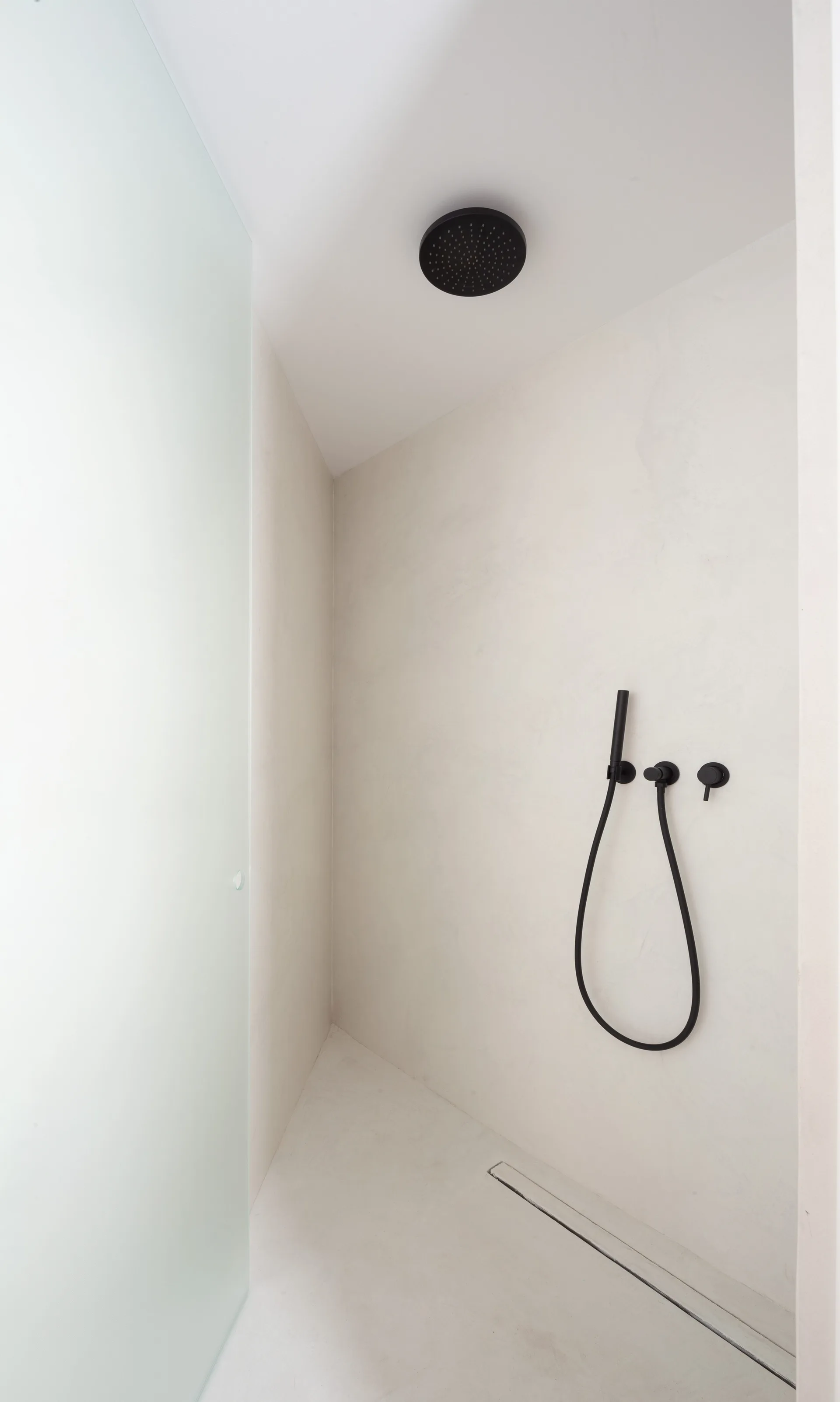 A minimalist walk-in shower features seamless off-white plaster walls, matte black fixtures, a frosted glass door, and a slim linear floor drain.