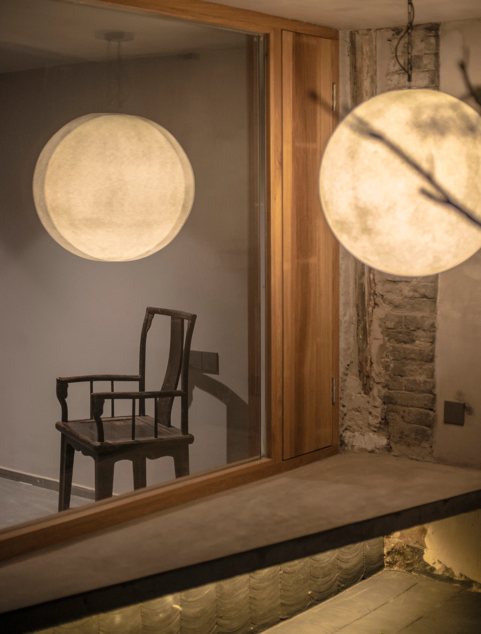 A glowing spherical pendant light hangs next to a glass partition showing the reflection of a dark wooden chair against an exposed brick wall.