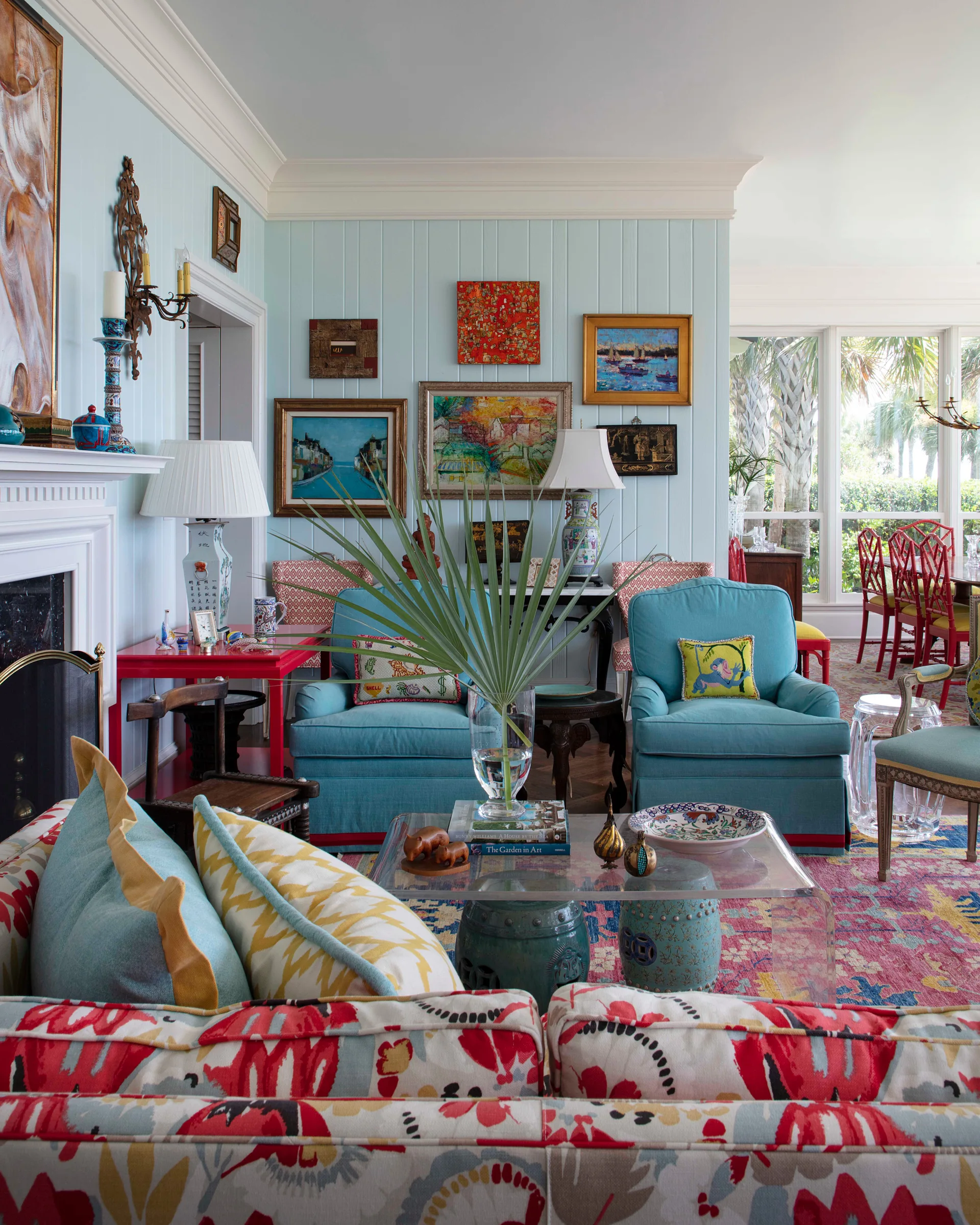 A colorful living room features a floral sofa, light blue armchairs, a clear acrylic coffee table, and art on light blue paneled walls.