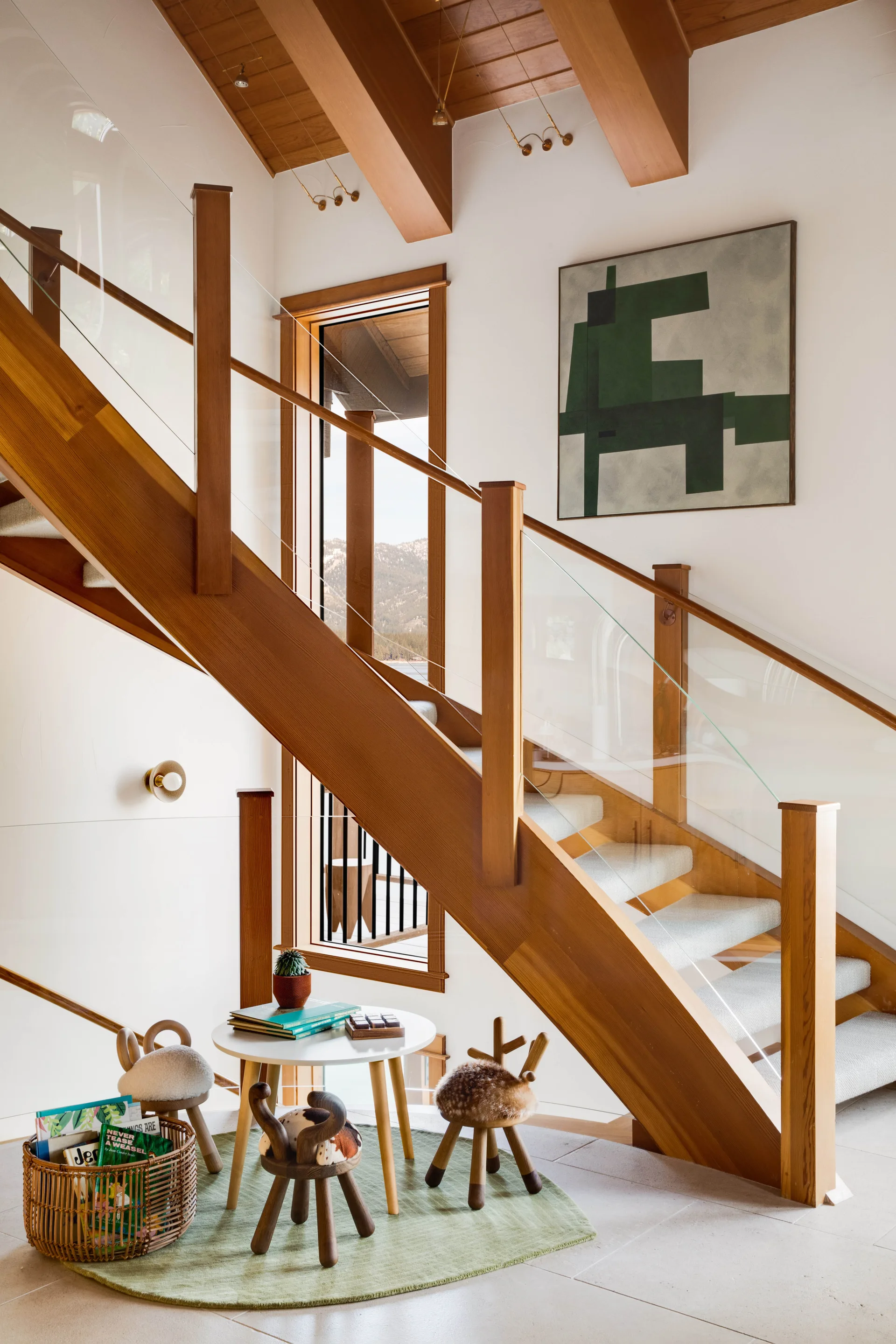 A wooden staircase with clear glass railings stands next to a small children's play area featuring animal-themed stools and a round white table.