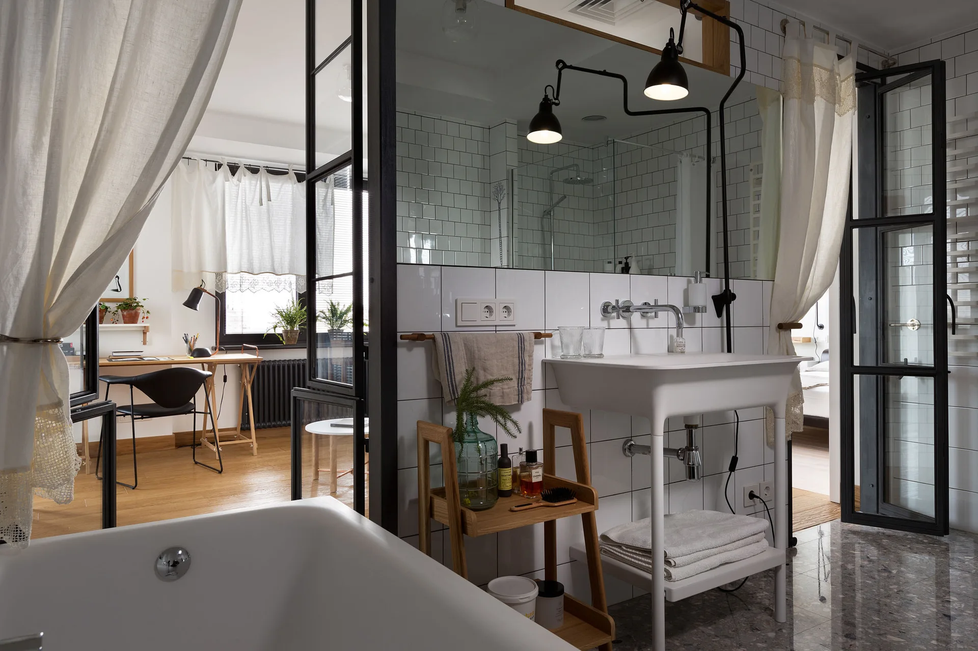 A modern bathroom features white subway tiled walls, a pedestal sink, and a black steel-framed glass partition looking into a bright home office.