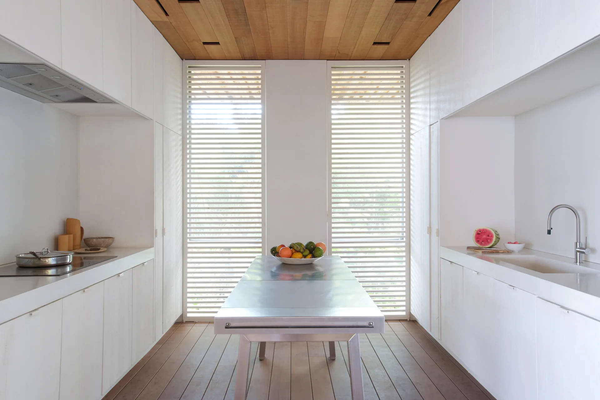 A minimalist galley kitchen featuring white cabinetry, a central stainless steel table, tall slatted windows, and a wood plank ceiling.