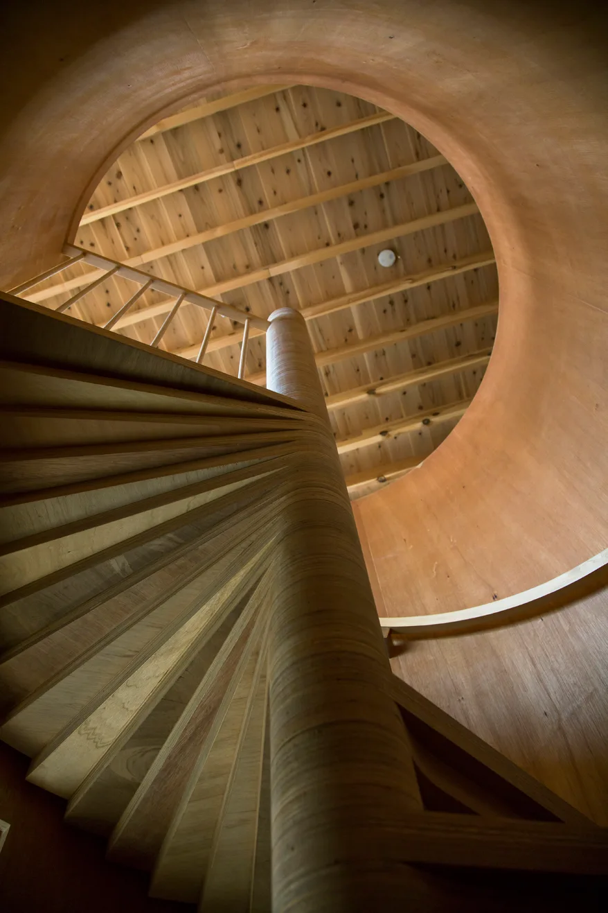 An upward view of a natural wood spiral staircase ascending through a smooth circular opening toward an exposed timber ceiling.