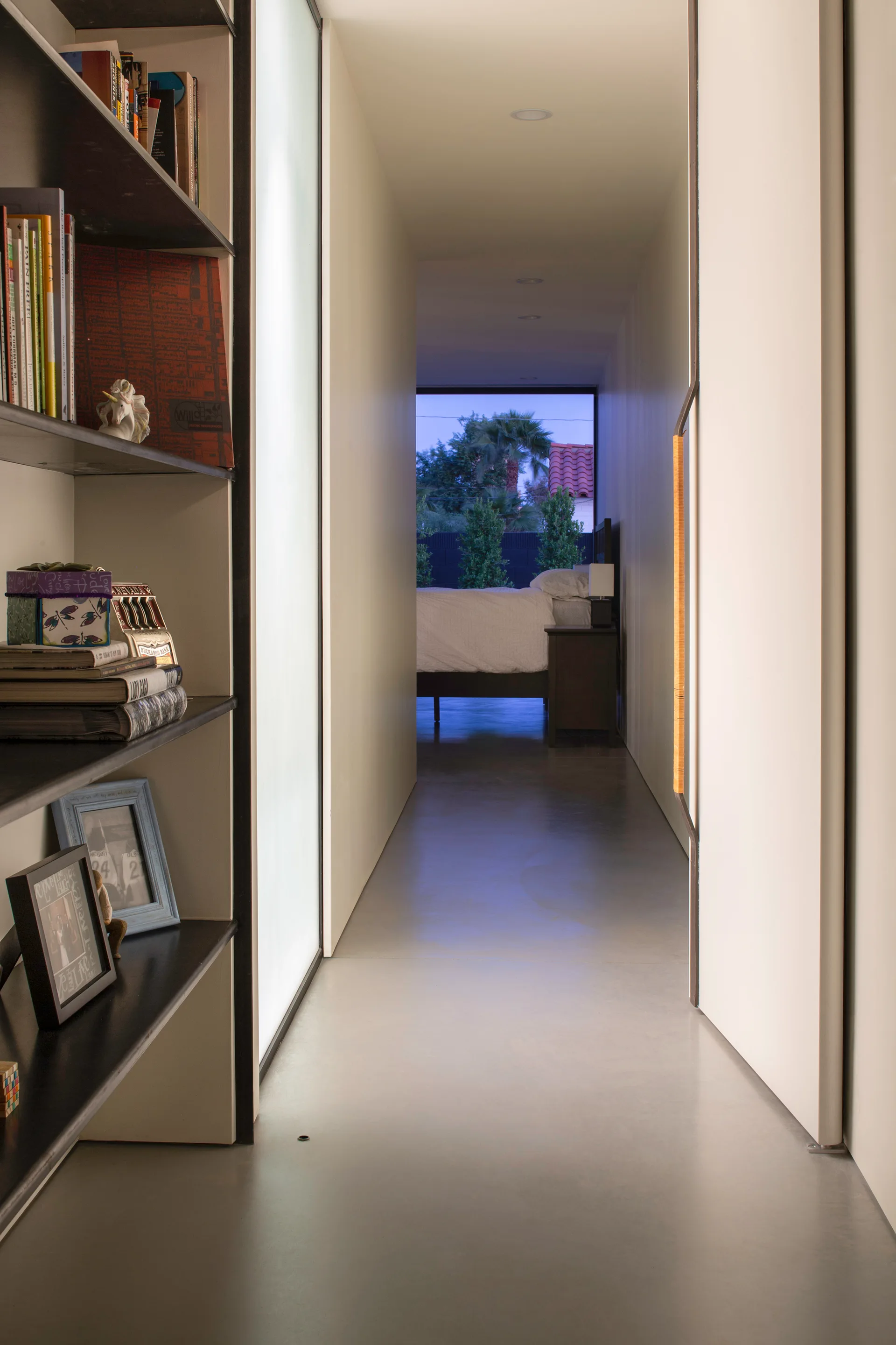 A minimalist hallway with smooth concrete flooring, a black built-in bookshelf, and a sliding white door leads toward a dimly lit bedroom.
