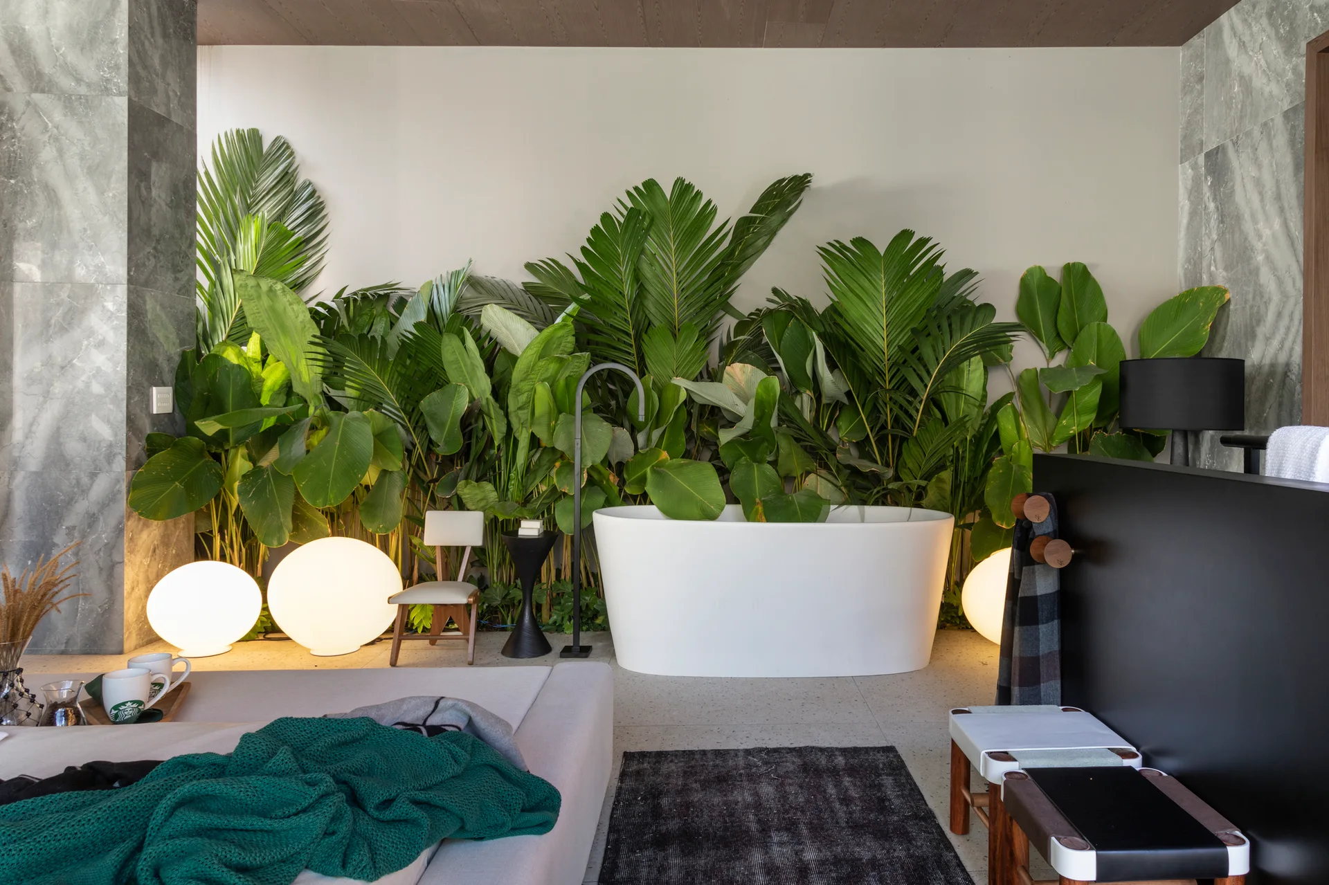 A modern bathroom features a white freestanding bathtub positioned in front of a wall of lush tropical plants and illuminated by glowing spherical floor lamps.