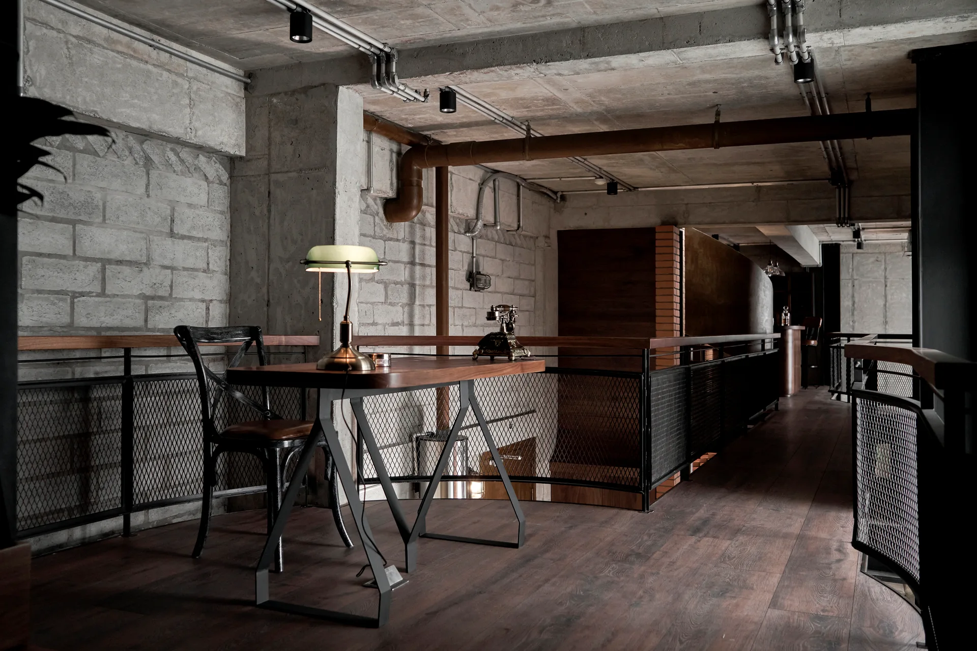 An industrial mezzanine workspace features a wood desk, vintage green bankers lamp, black metal mesh railing, and raw concrete walls.