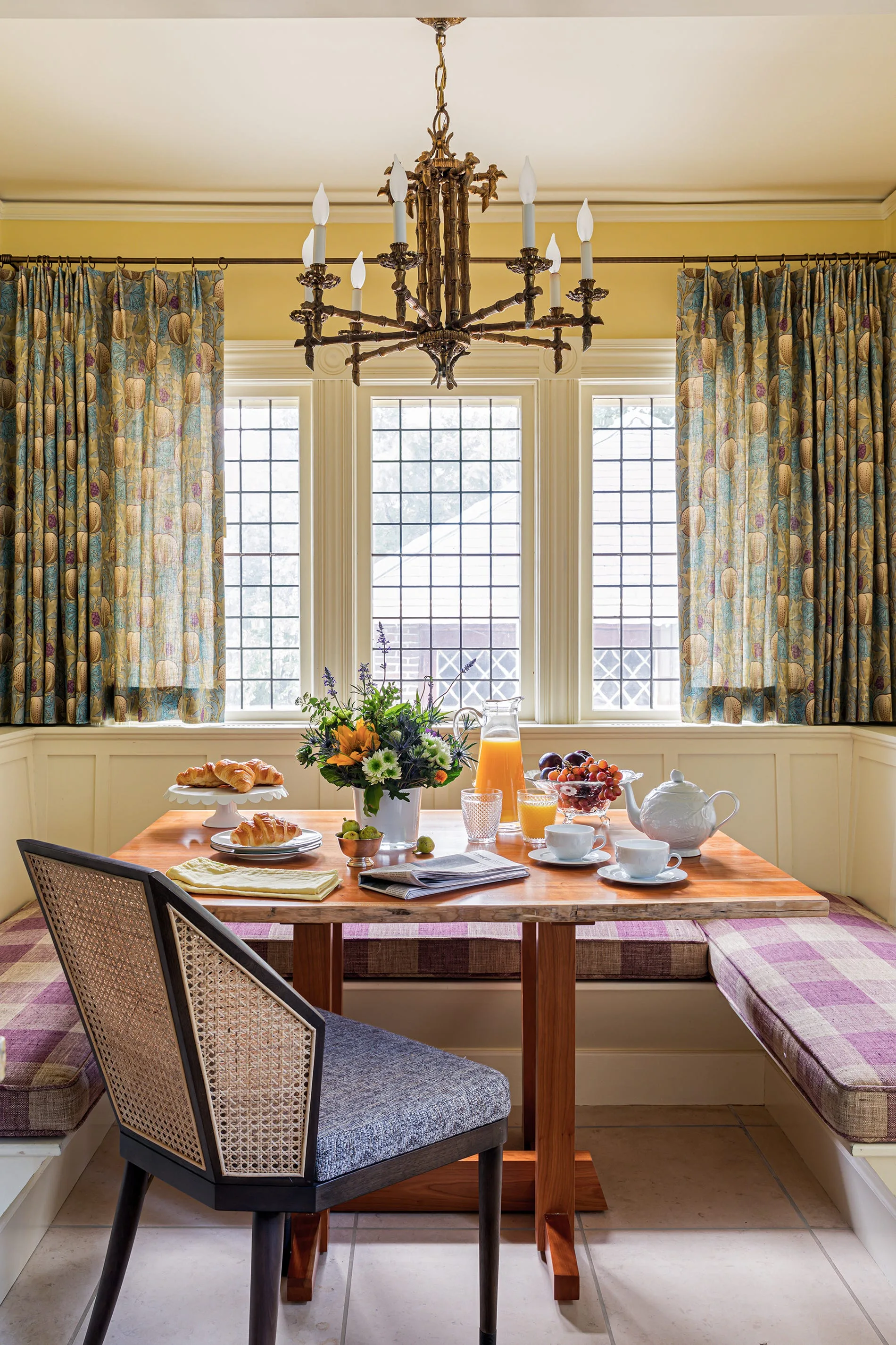A sunlit breakfast nook features a wooden dining table, a built-in banquette with purple plaid cushions, and a cane-back chair beneath a metal chandelier.
