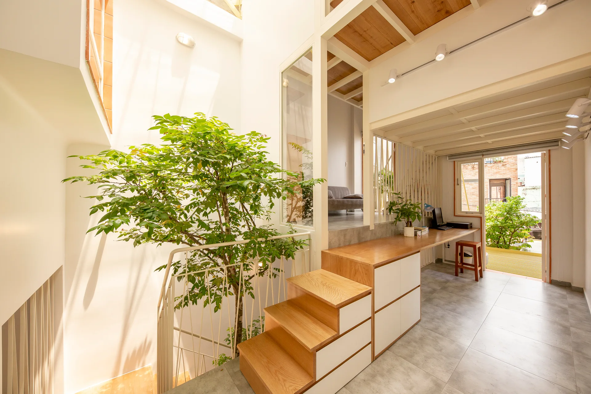 A multi-level interior showing a light wood staircase with built-in white storage drawers, an adjacent desk, and a green indoor tree.