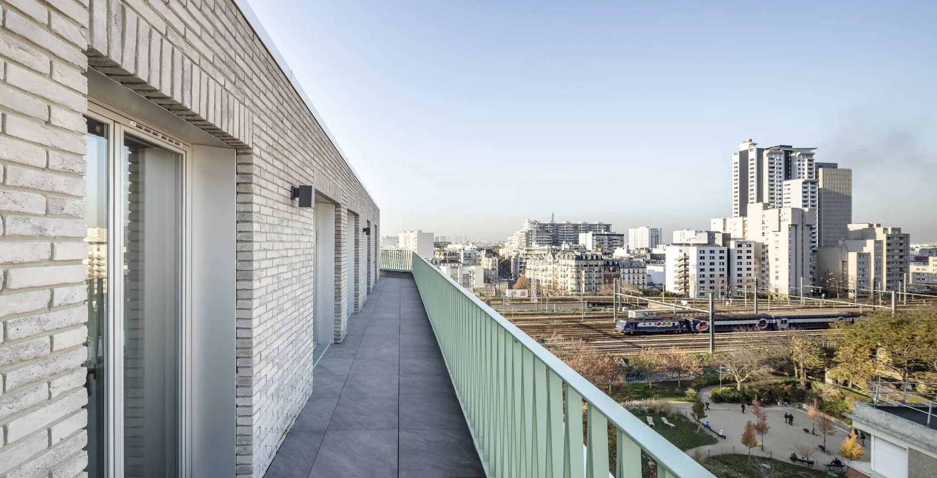 A long outdoor balcony featuring light grey brick walls, grey floor tiles, and a light green metal railing overlooking a cityscape.