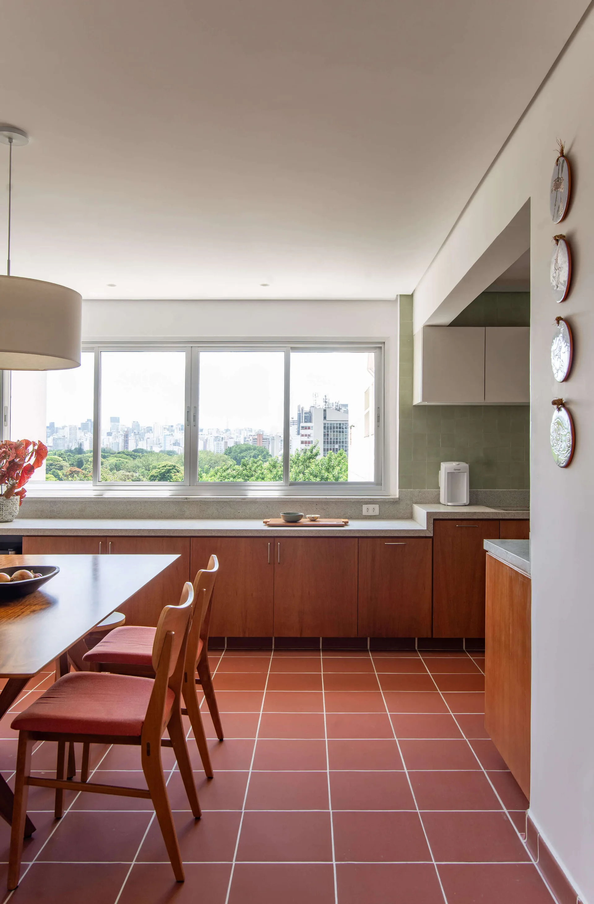 A dining area featuring a wood table and chairs with terracotta floor tiles and a long kitchen counter beneath a large window showing a city view.