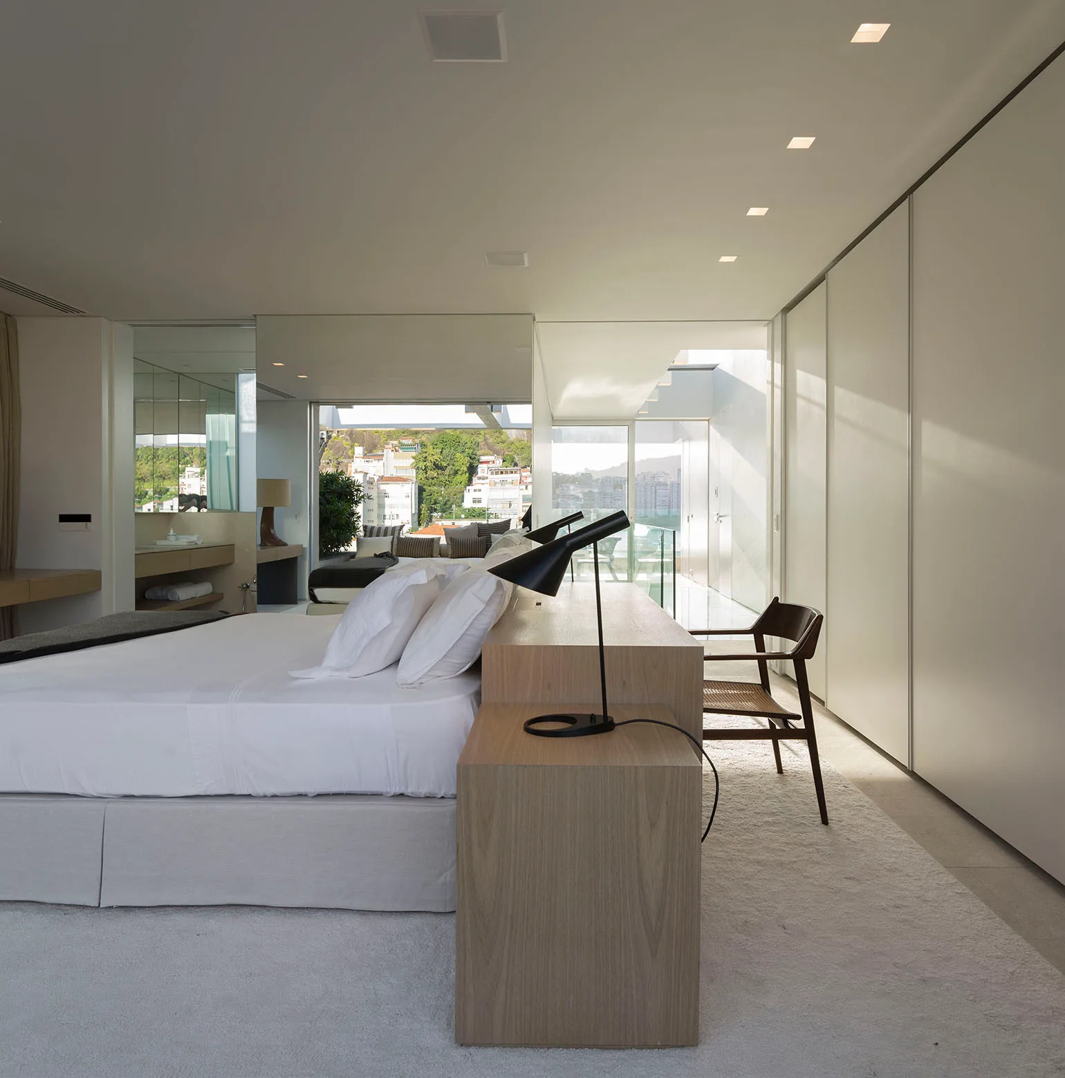 A minimalist white bedroom featuring a central bed backed by a light wood console table with a black desk lamp and dark wooden chair.