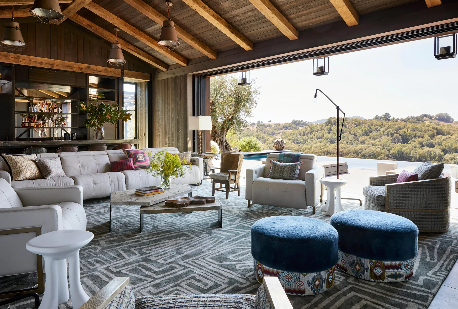 A rustic living room features a cream sectional, blue ottomans, and exposed wooden beams, opening completely to an outdoor pool terrace.
