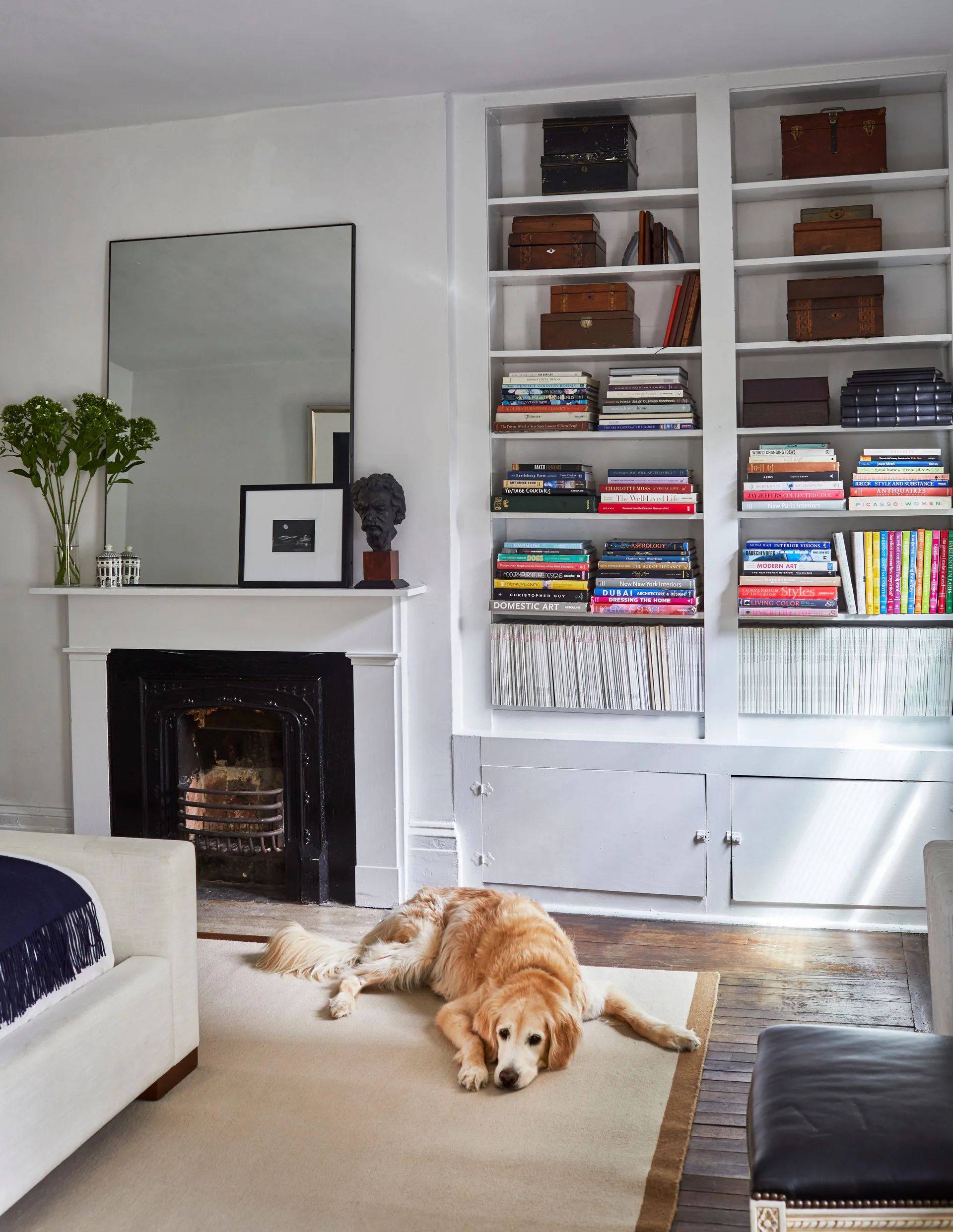 A living room features white built-in bookshelves filled with books next to a white fireplace with a black insert and a large mirror.