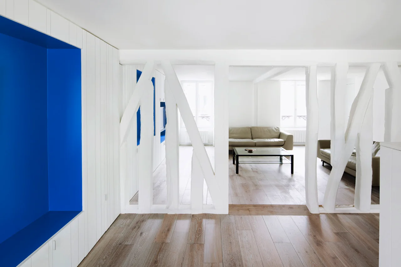 A white painted timber partition divides a hallway with a cobalt blue recessed niche from a bright living room featuring a leather sofa and glass table.