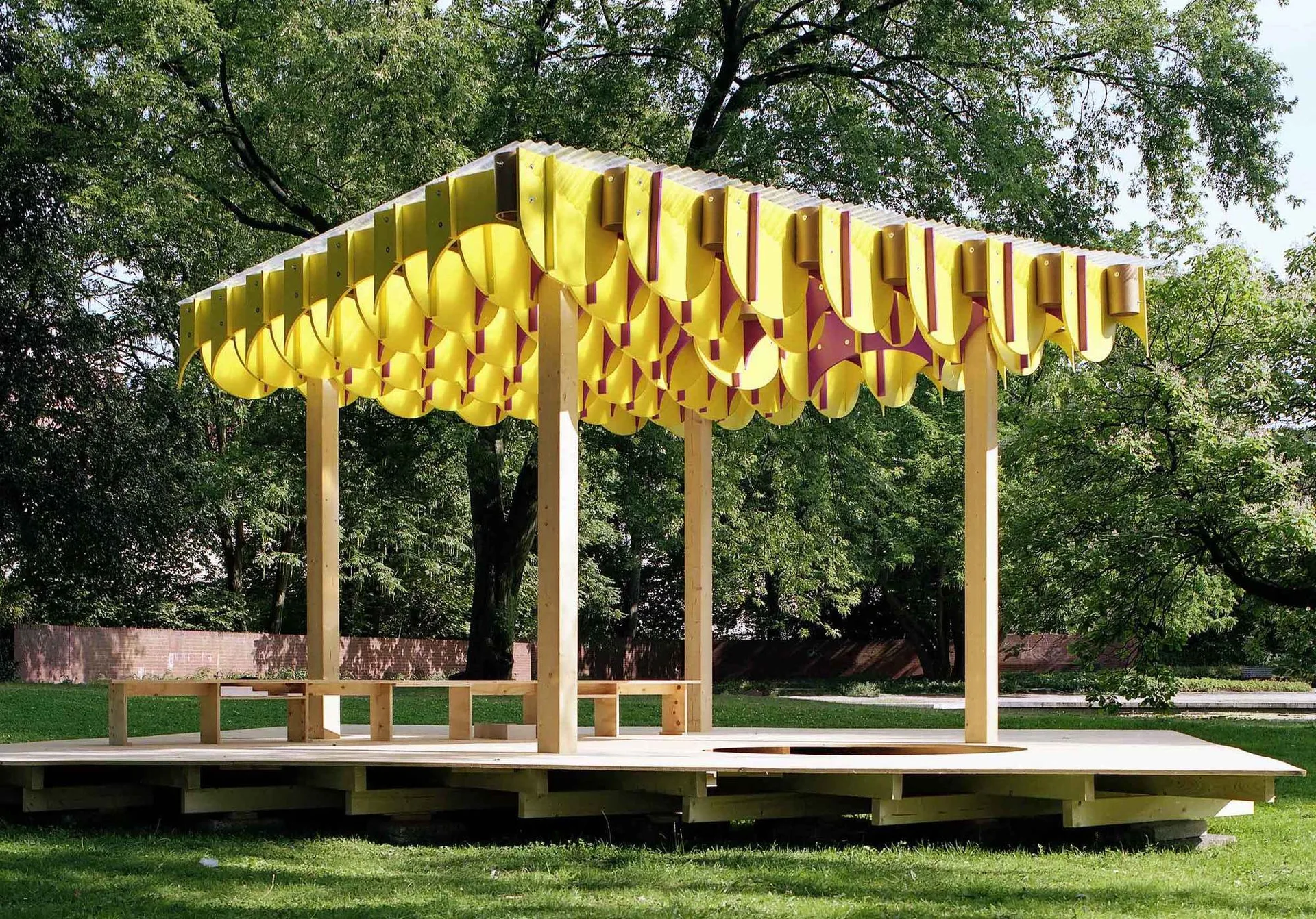 A raised wooden pavilion with simple benches features a unique, scalloped yellow and purple fabric canopy roof in a green park setting.