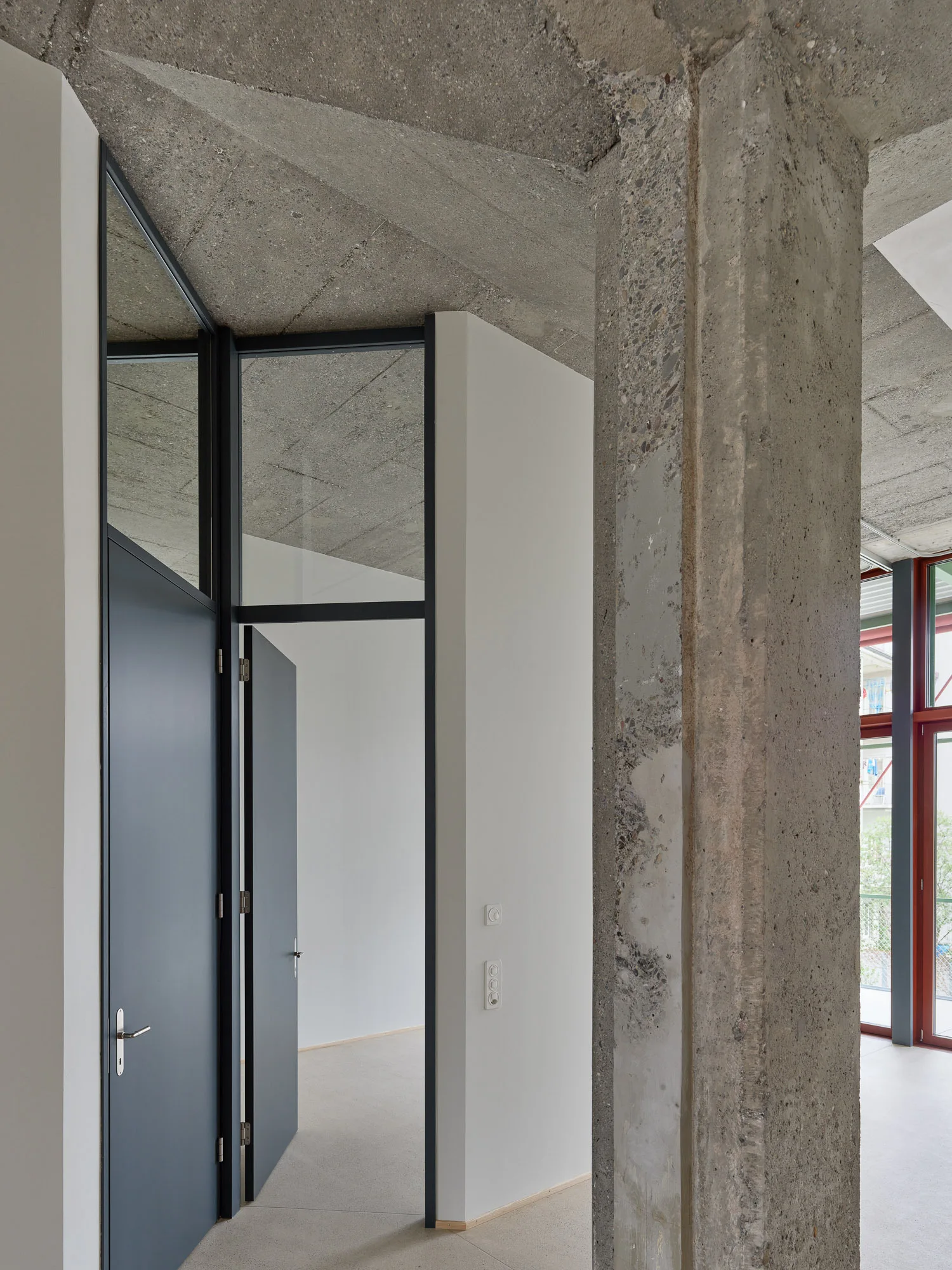 An interior hallway features a thick, raw concrete column and an exposed concrete ceiling contrasted against smooth white walls and dark grey doors.