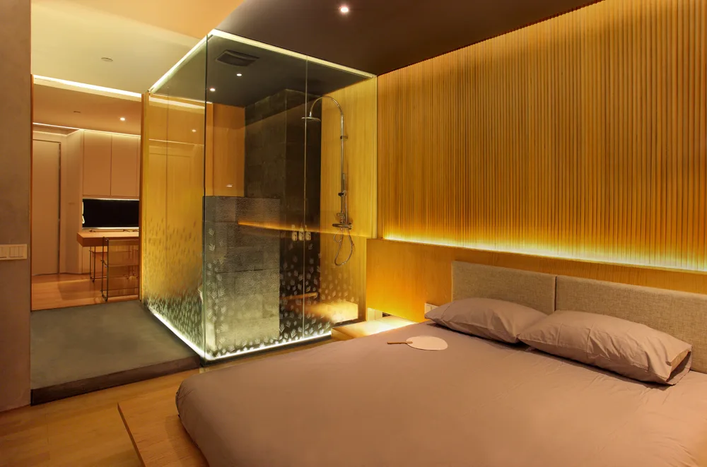 A modern bedroom features a low platform bed alongside an integrated glass-enclosed shower, highlighted by a vertical wood-slatted wall with warm lighting.