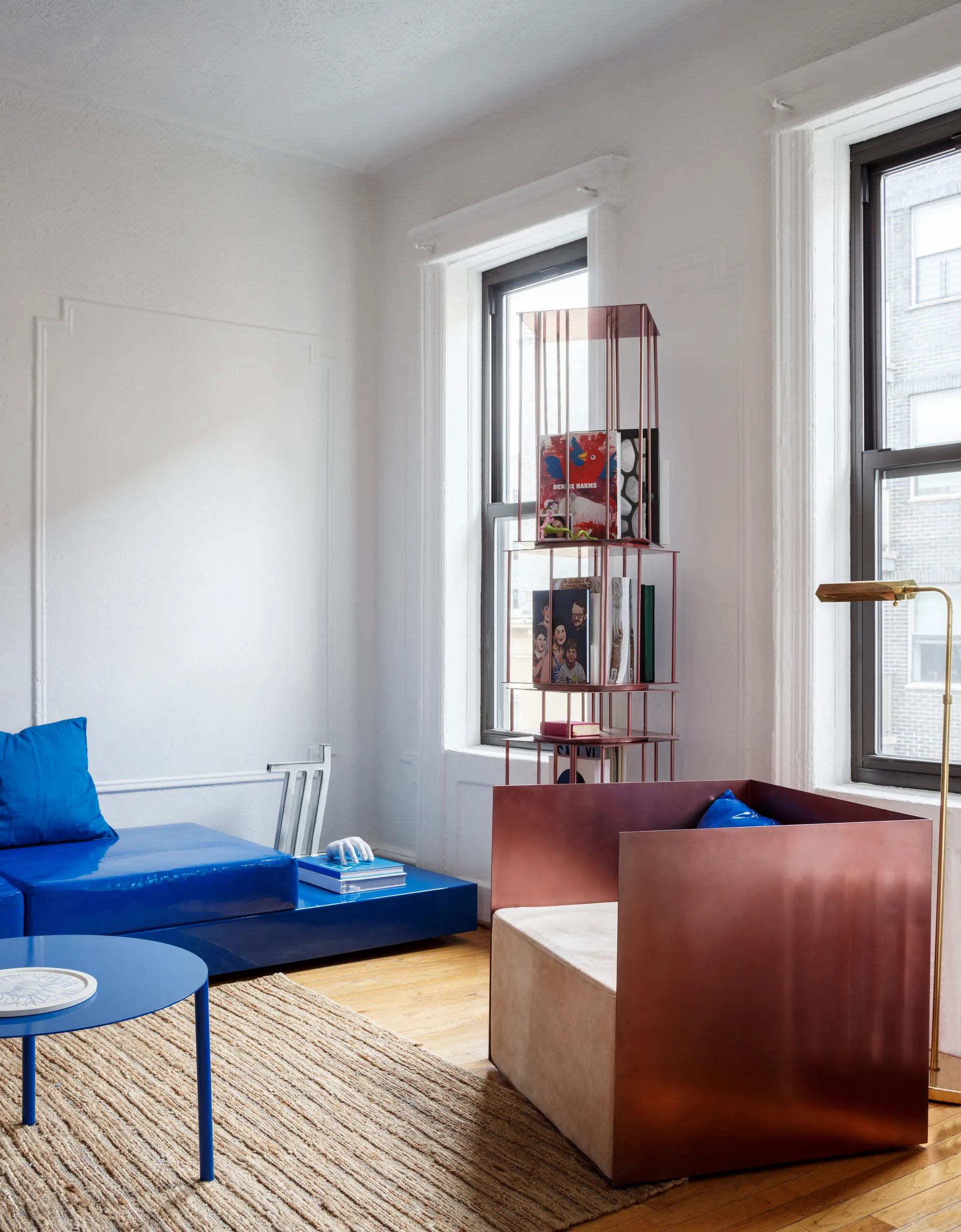 A modern living room features a glossy blue modular sofa and a metallic copper armchair on a woven jute rug next to two windows.