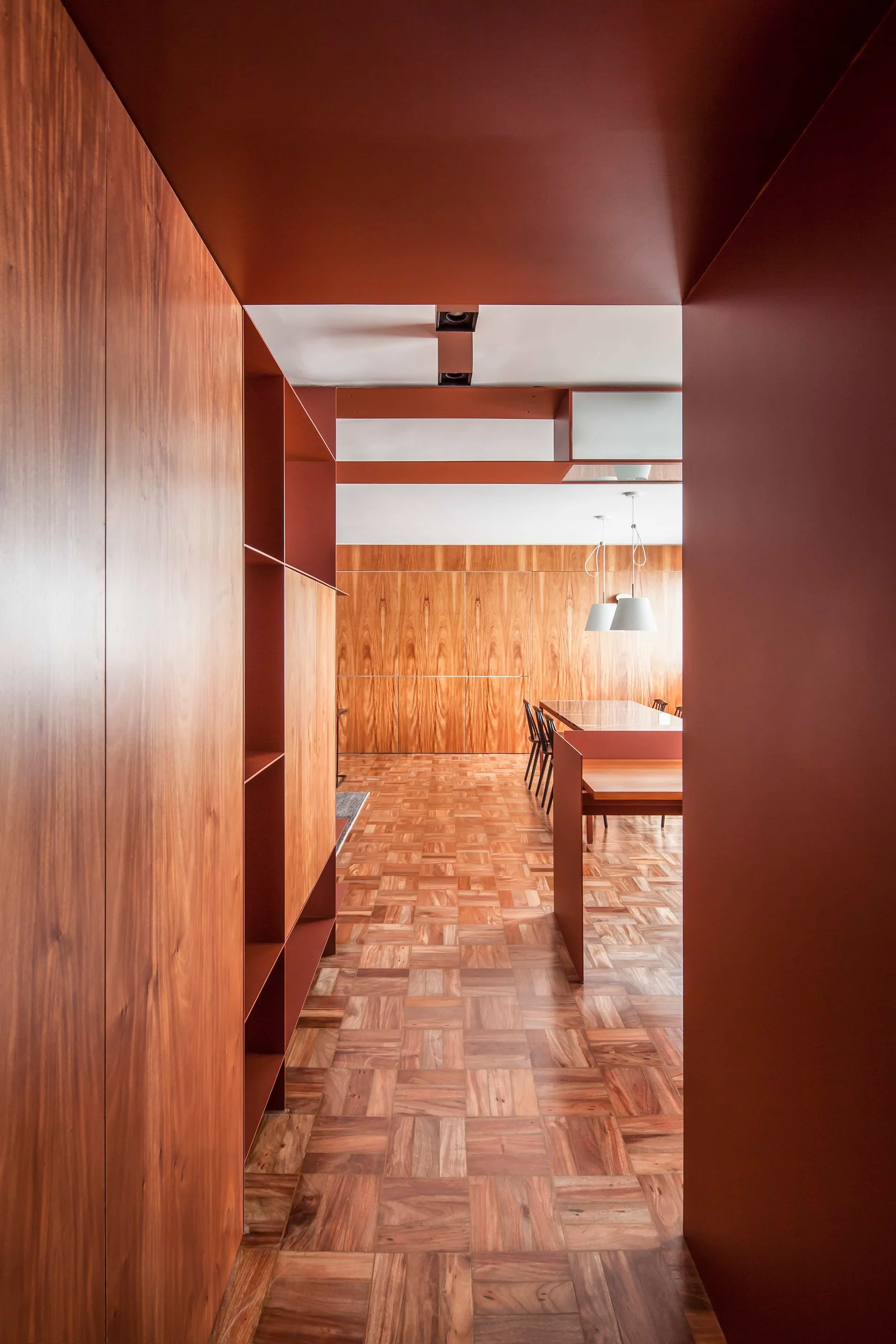 A modern dining area with wood-paneled walls and a parquet floor is viewed through a matte rust-colored architectural entryway.