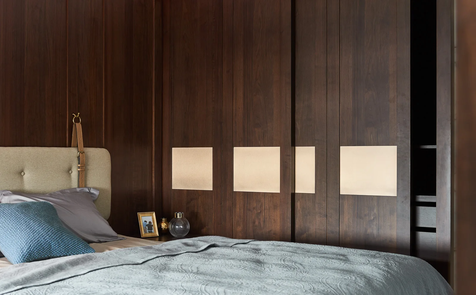 A bedroom bed sits beside dark wood paneled walls and built-in sliding wardrobe doors with inlaid square brass panels.