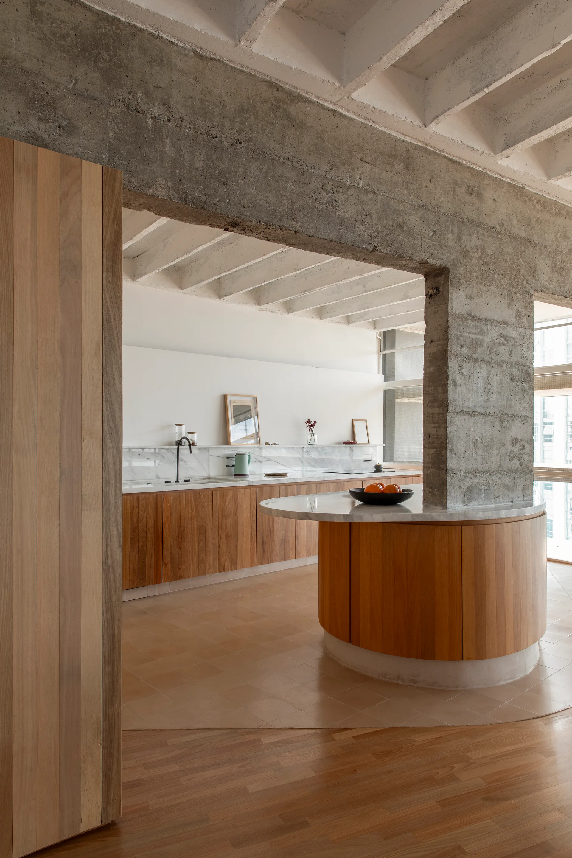 A modern kitchen featuring a curved wood-toned island with a marble top under a raw concrete ceiling and structural columns.