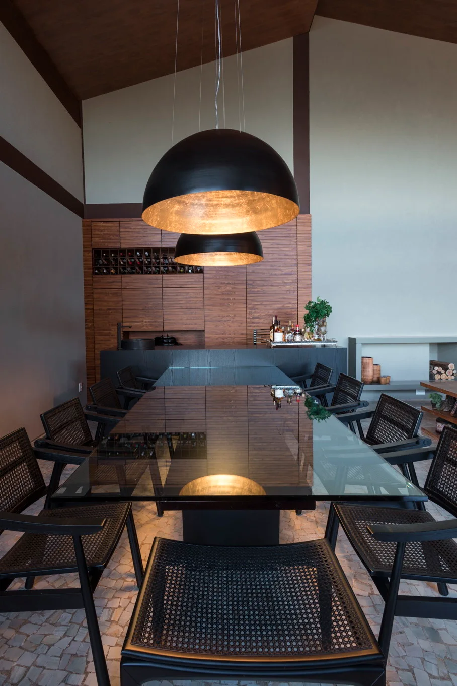 A modern dining room features a long glass-topped table surrounded by black mesh chairs under two large black dome pendant lights with gold interiors.