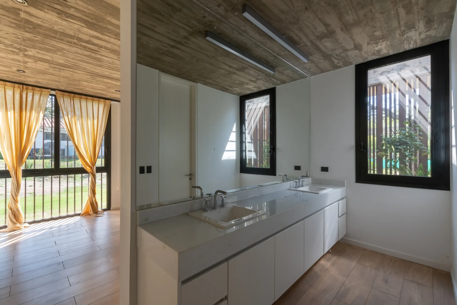 A modern bathroom features a white double vanity with dual sinks, a large wall mirror, and an exposed raw concrete ceiling above wood floors.