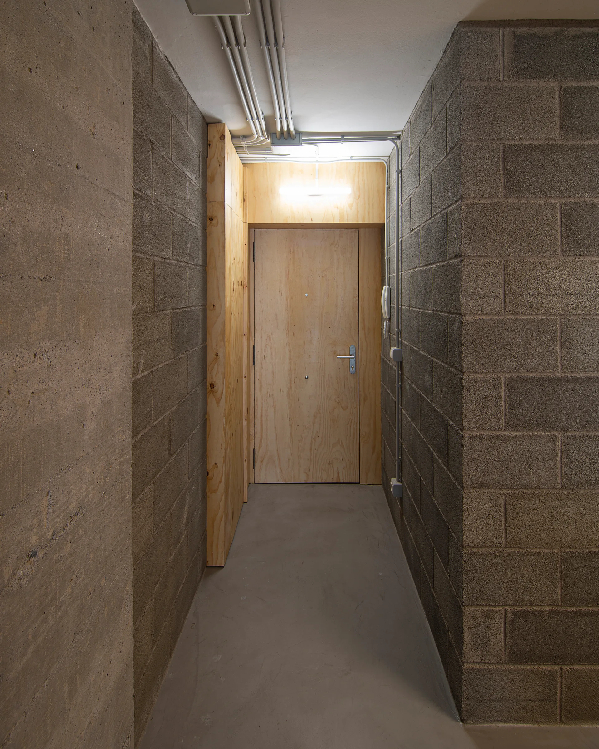 A narrow concrete hallway features raw cinder block walls, a smooth grey floor, and a natural plywood door at the end under exposed metal conduit pipes.