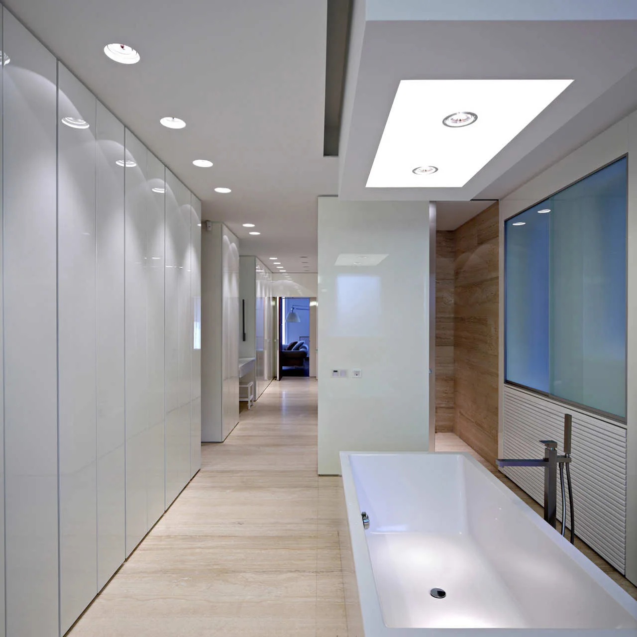A modern minimalist bathroom features a white rectangular bathtub, high-gloss white floor-to-ceiling cabinets, and polished beige stone tile flooring.